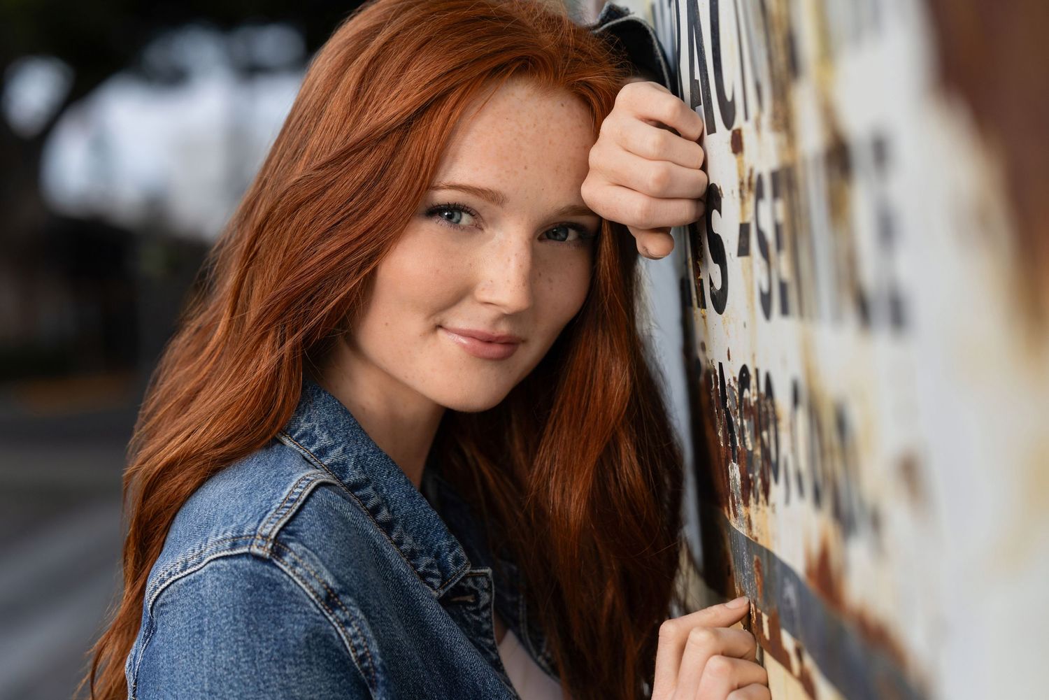 Young woman with long red hair leaning against a graffiti wall outdoors, wearing a denim jacket.