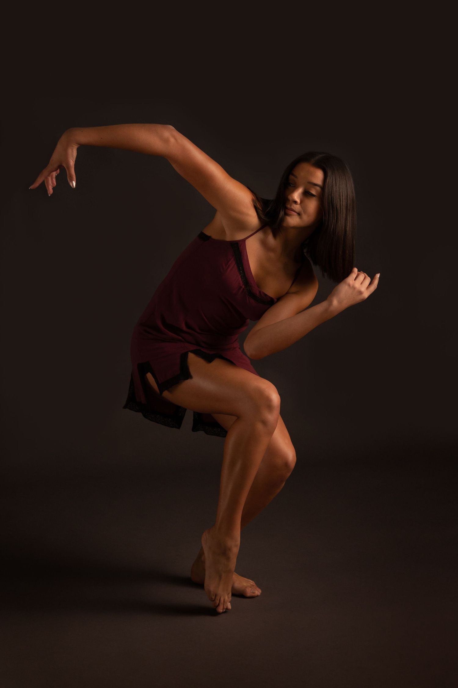 Female dancer in a burgundy dress performing a dynamic pose against a dark studio background.