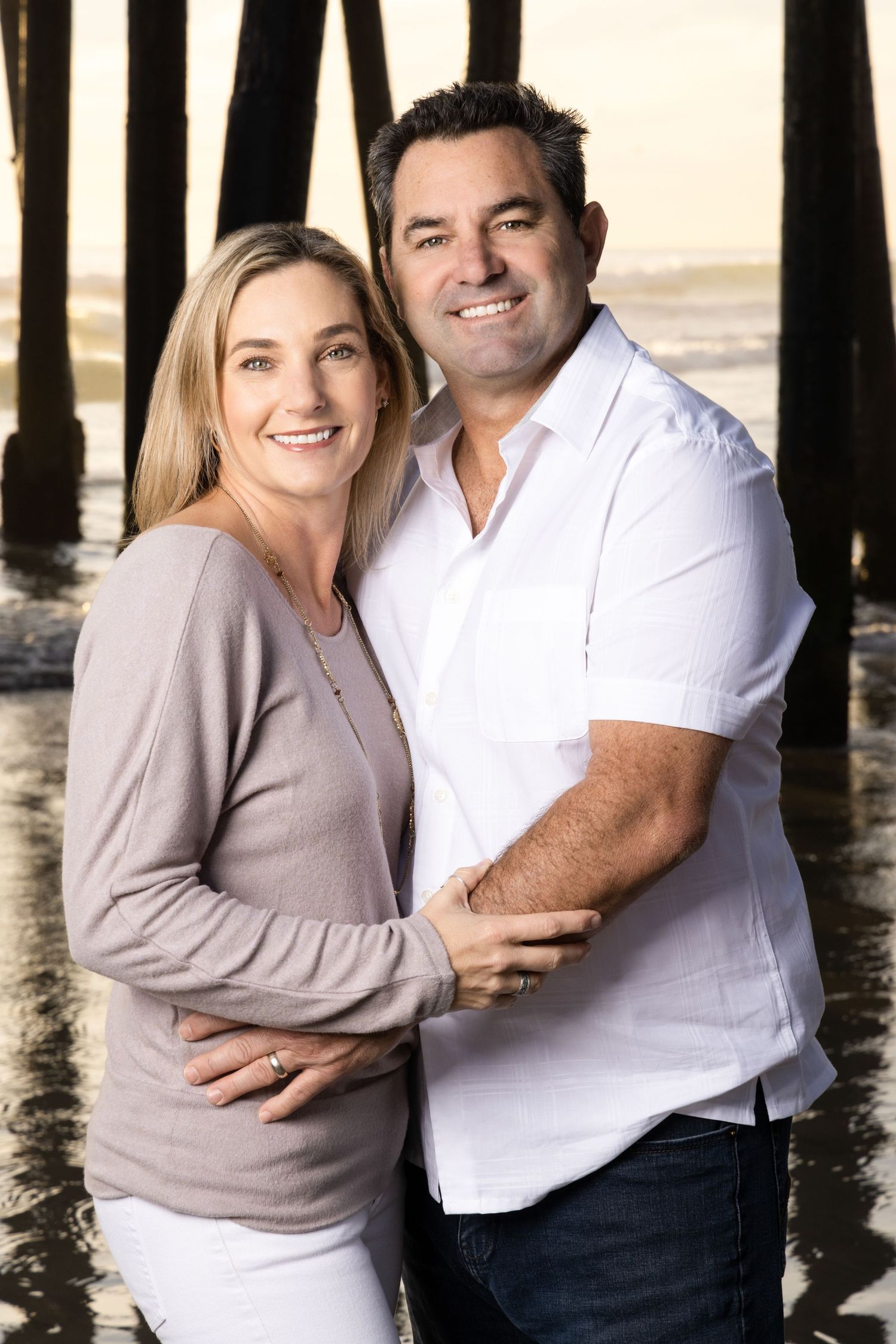 Smiling couple embracing under a pier at the beach during golden hour sunset.