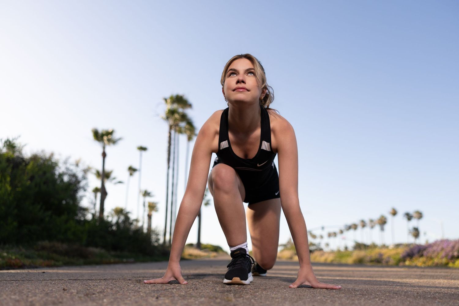 Woman in athletic wear in a runner's starting position on a road with palm trees in the background.