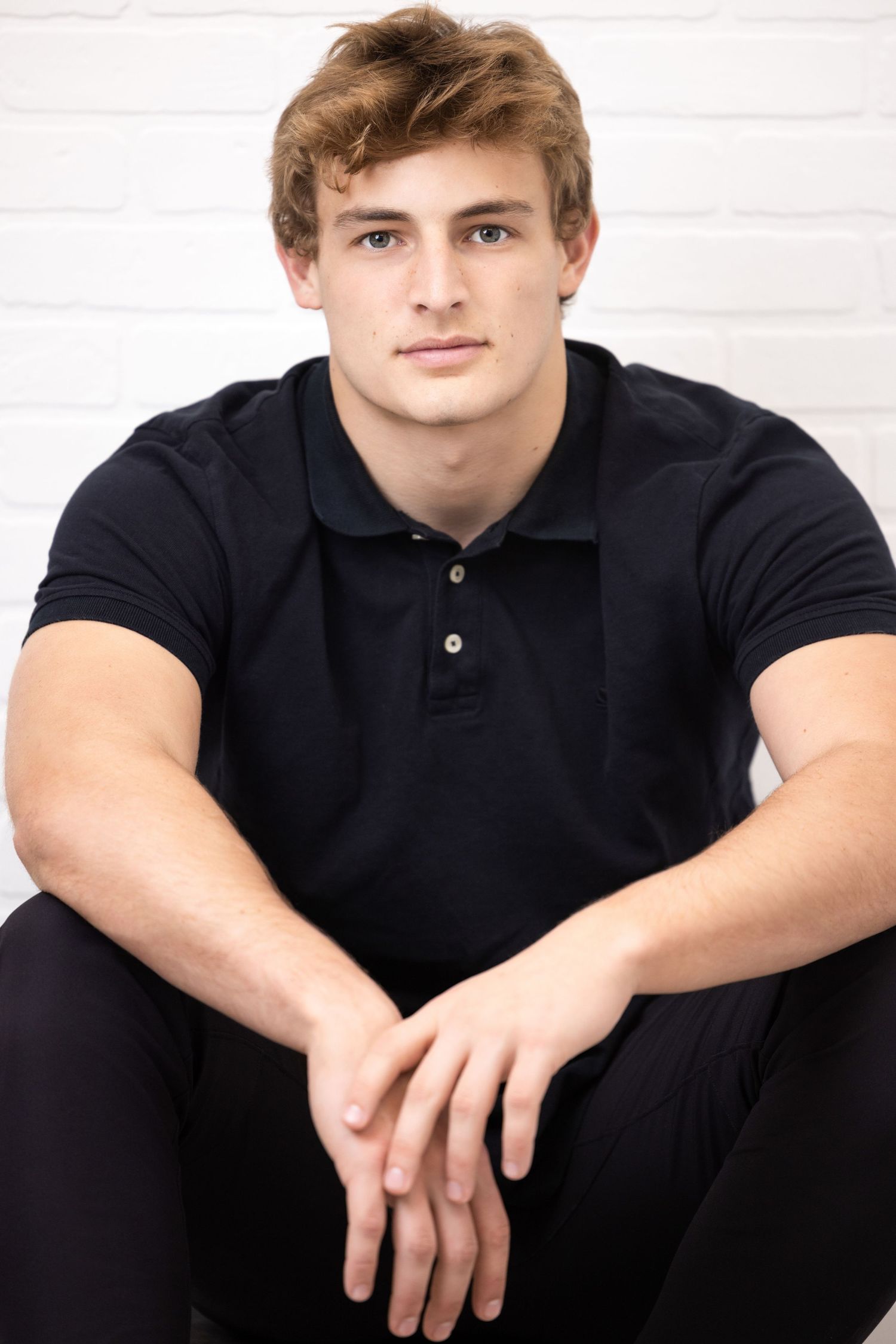 Athletic young man with blonde hair wearing a black polo shirt sitting against a white brick wall.