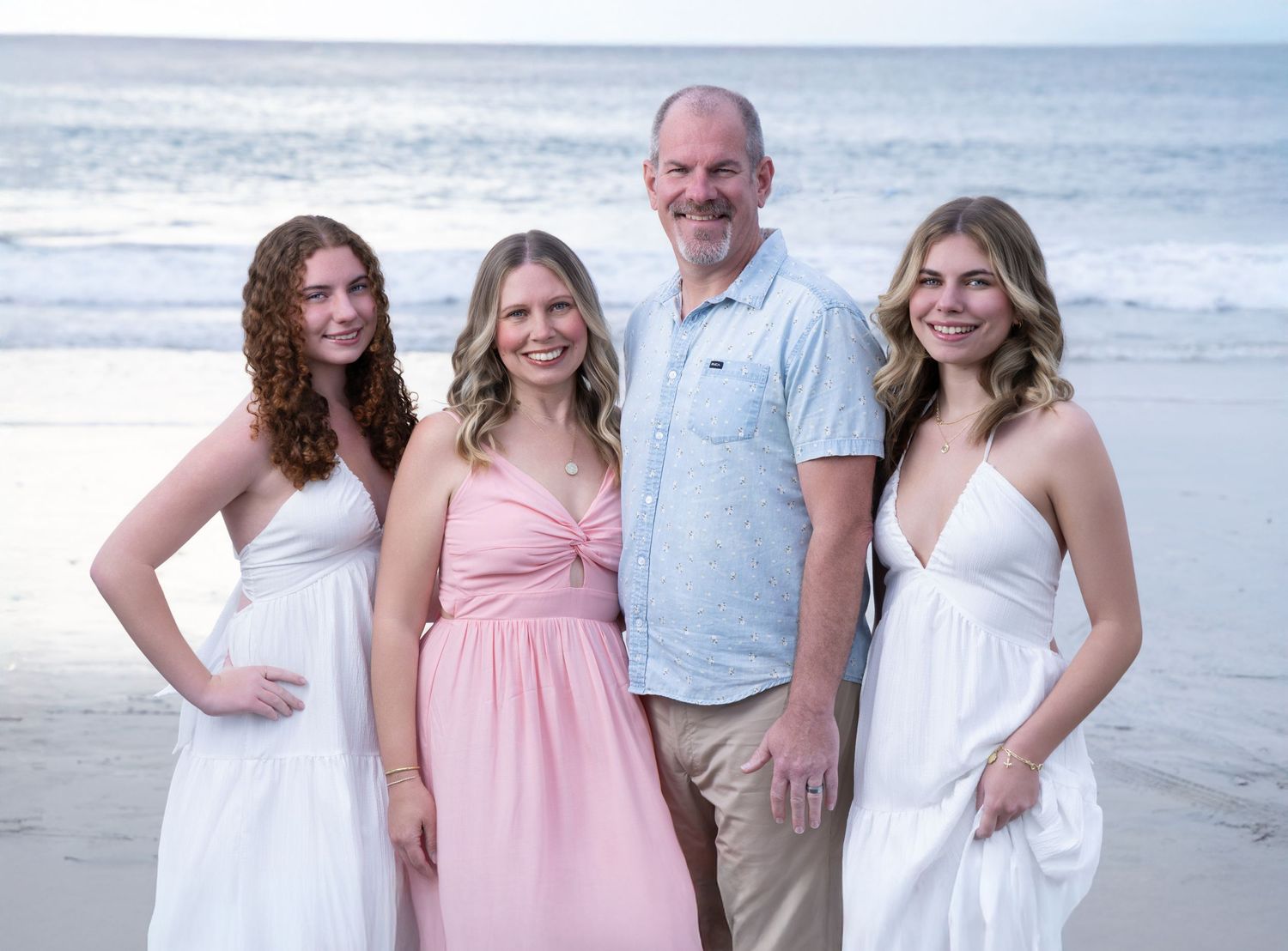 Family of four smiling together on a beach at sunset, wearing white and pink outfits.
