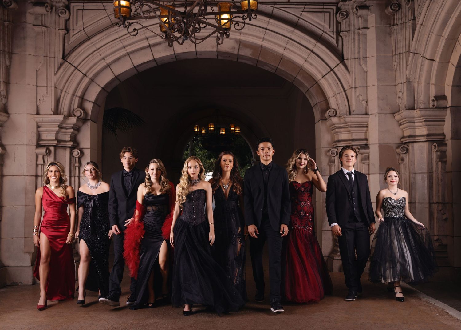 Group of elegantly dressed young adults in red and black formal wear pose under stone archway.
