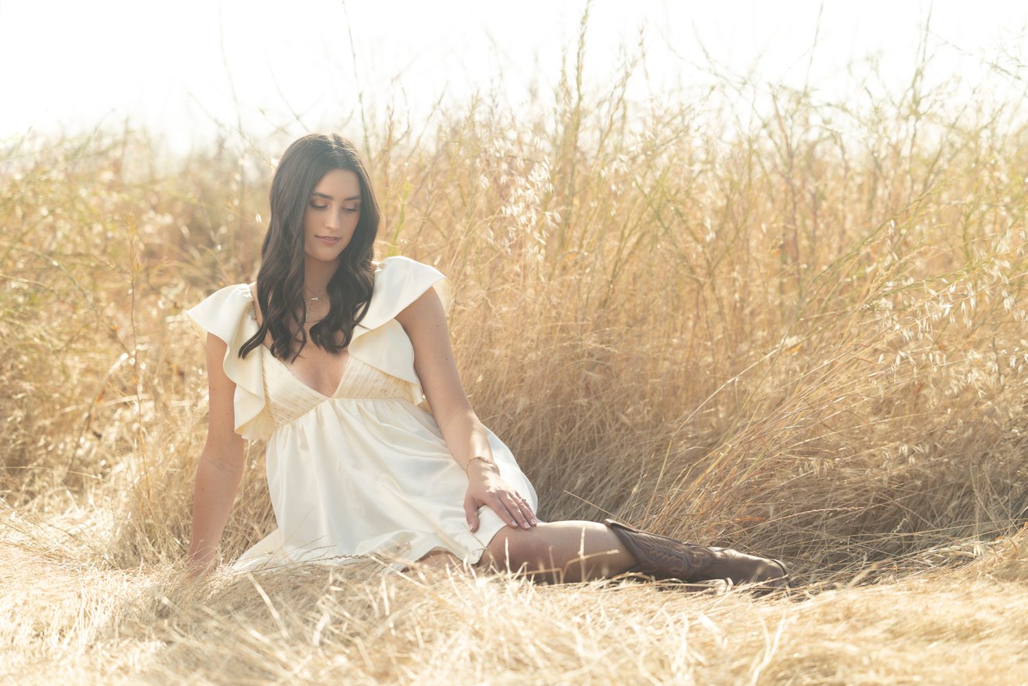 Young woman in white dress sitting gracefully in golden wheat field on sunny day.