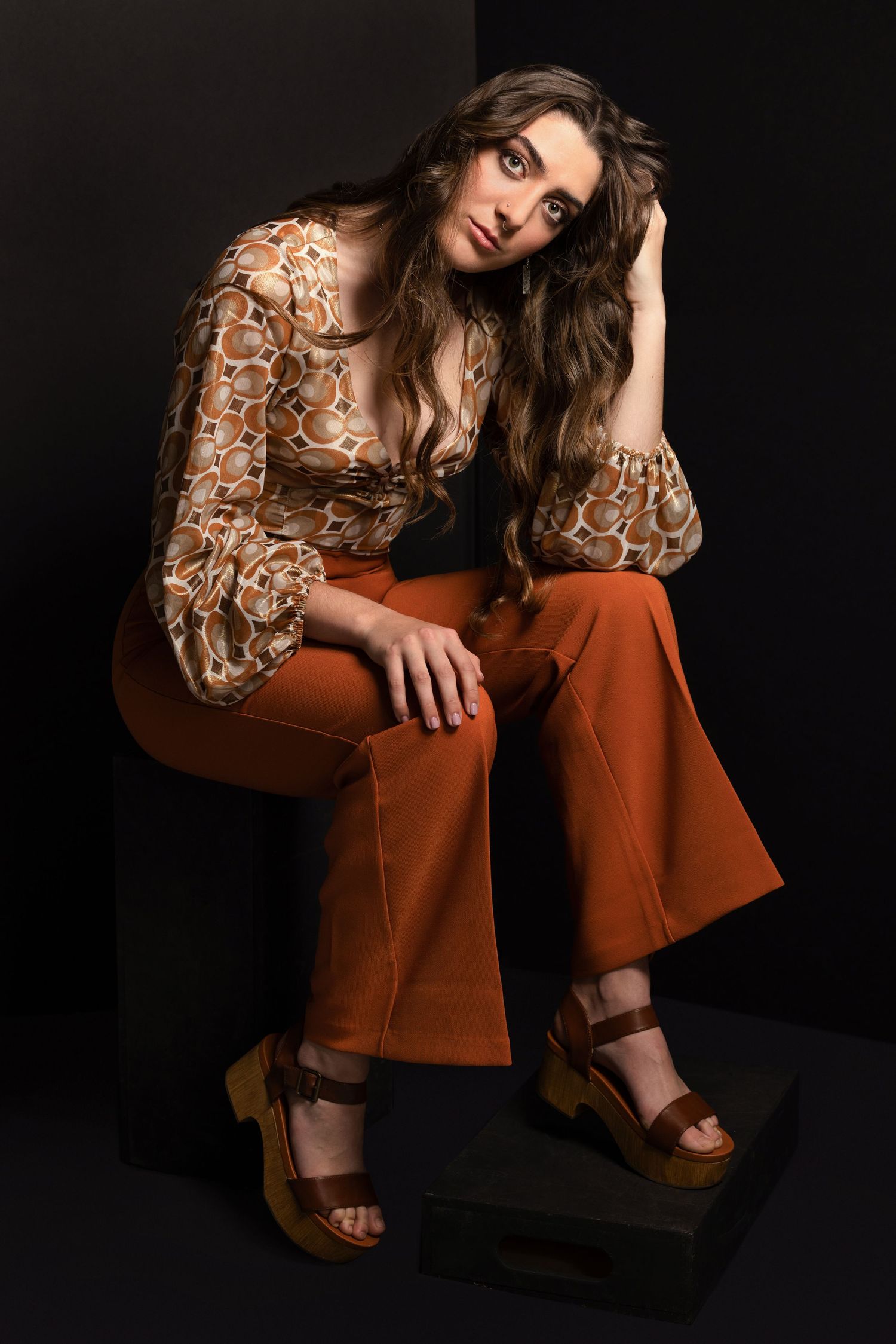 Woman in brown patterned blouse and rust wide-leg pants sits on black cube wearing tan platform sandals.