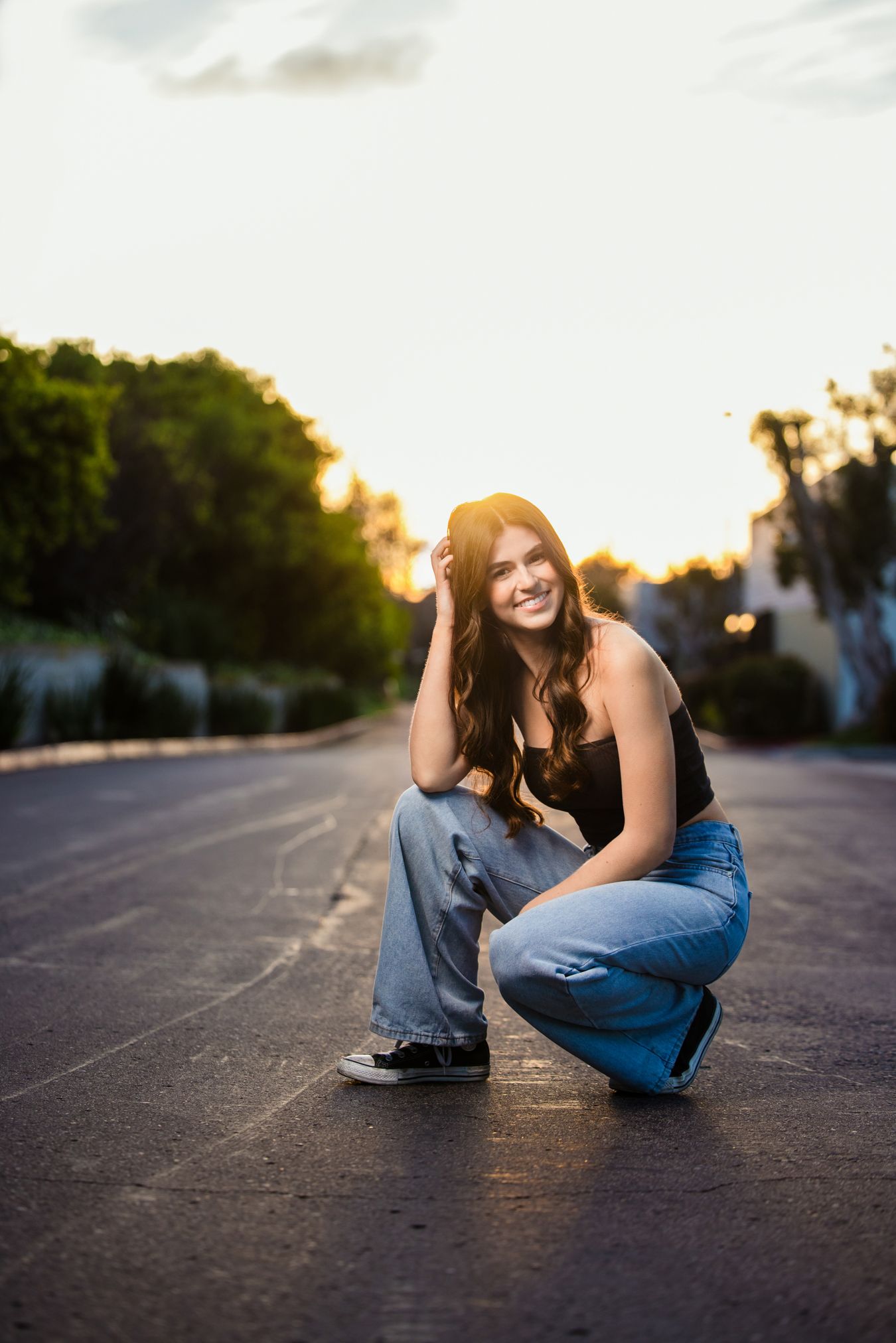 Young woman in jeans and black top crouching on a road at golden hour, smiling with long wavy hair.