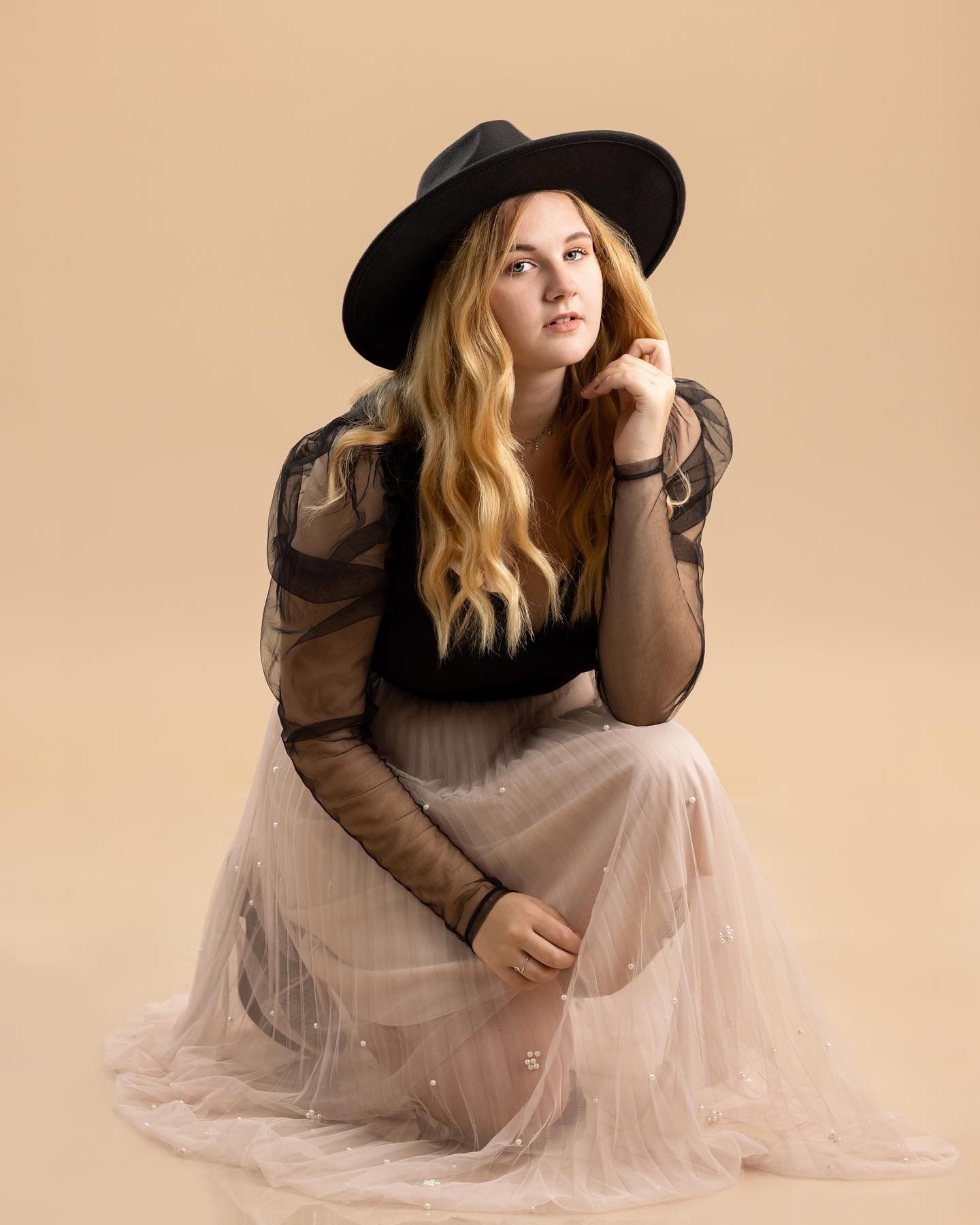 Young woman in black sheer top, wide-brim hat, and blush tulle skirt poses gracefully against a warm beige background.
