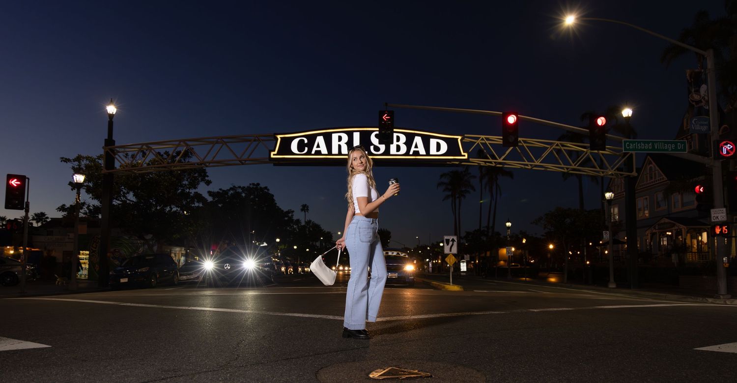 Woman in white top and jeans stands in the street beneath the illuminated Carlsbad village sign at dusk.