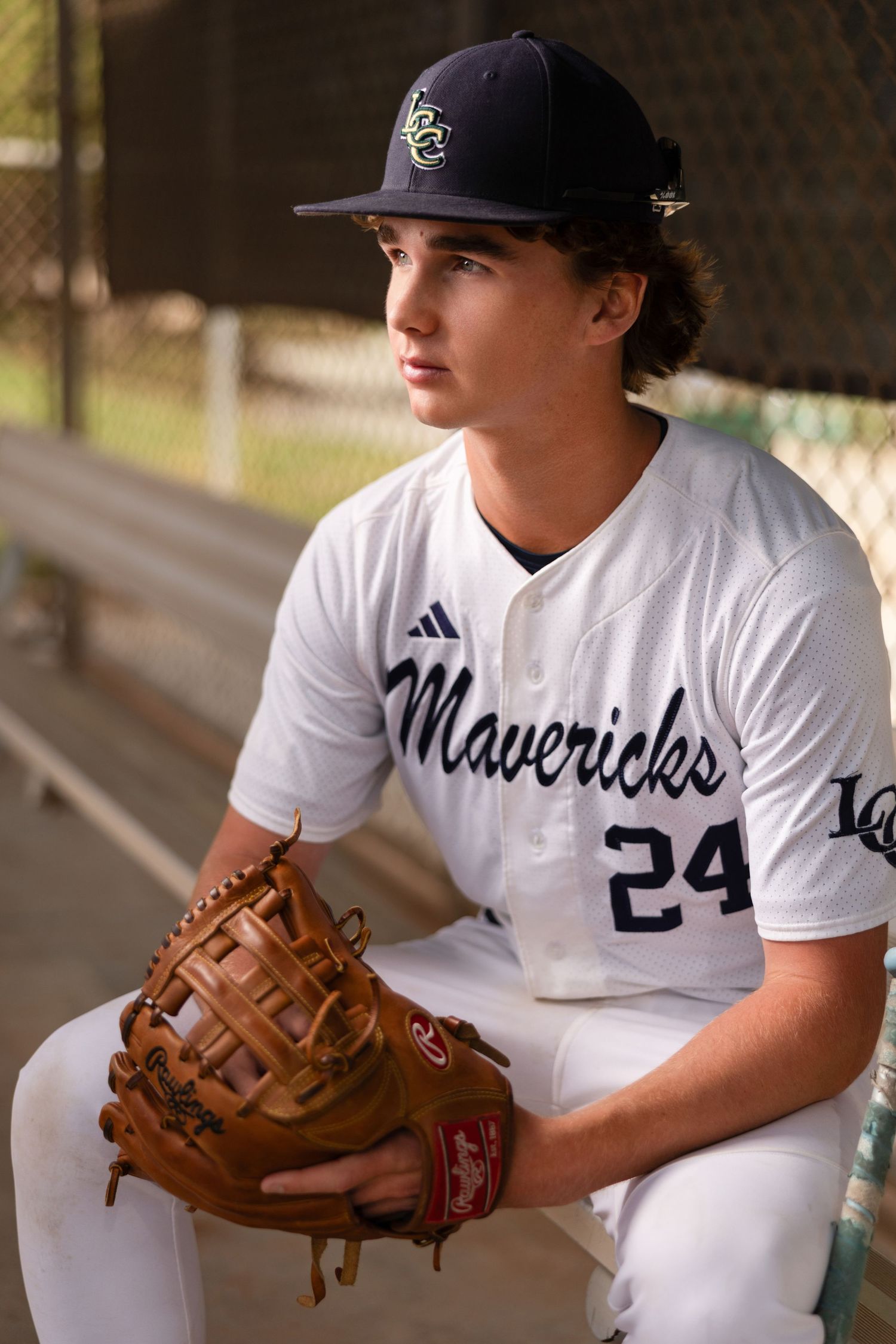 Young baseball player in Mavericks #24 jersey sits in dugout holding Rawlings glove, wearing team cap.