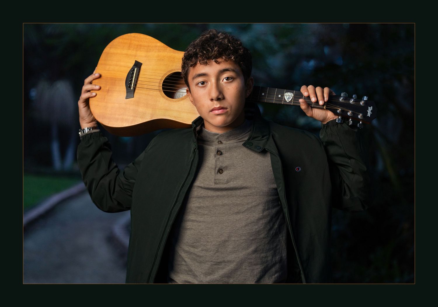 Young man holding acoustic guitar on his shoulder, wearing a dark jacket, posing outdoors for a music artist portrait.