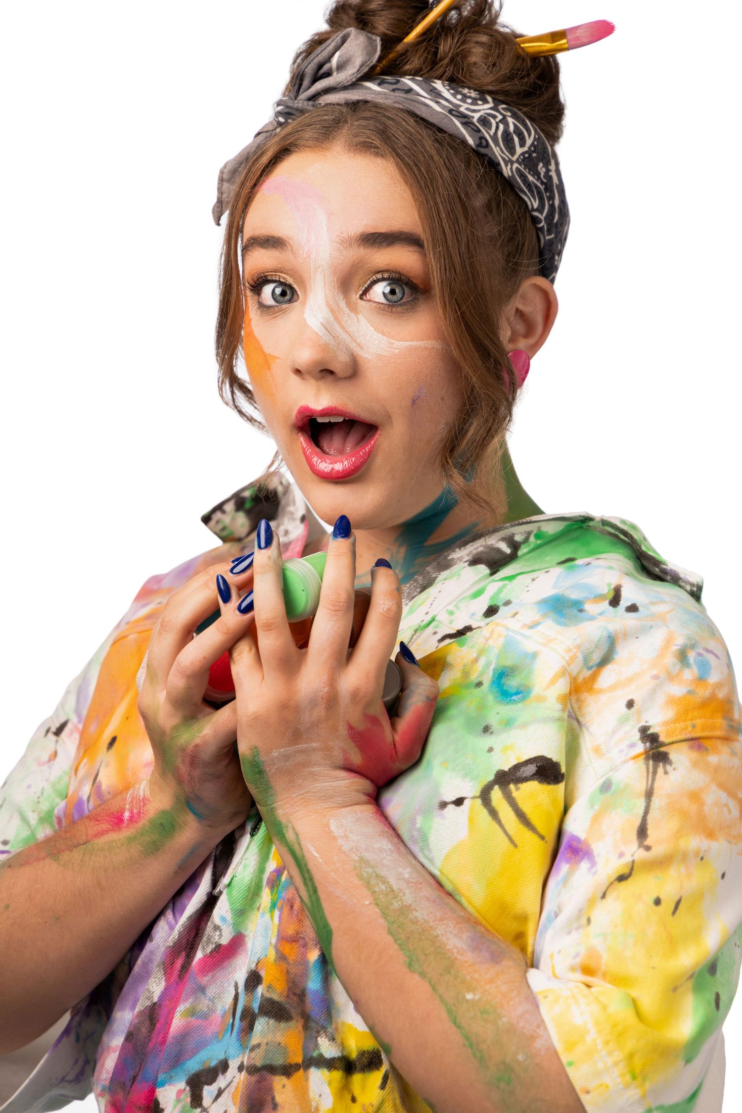 Young woman covered in colorful paint splatters with brushes in hair, looking surprised against white background.