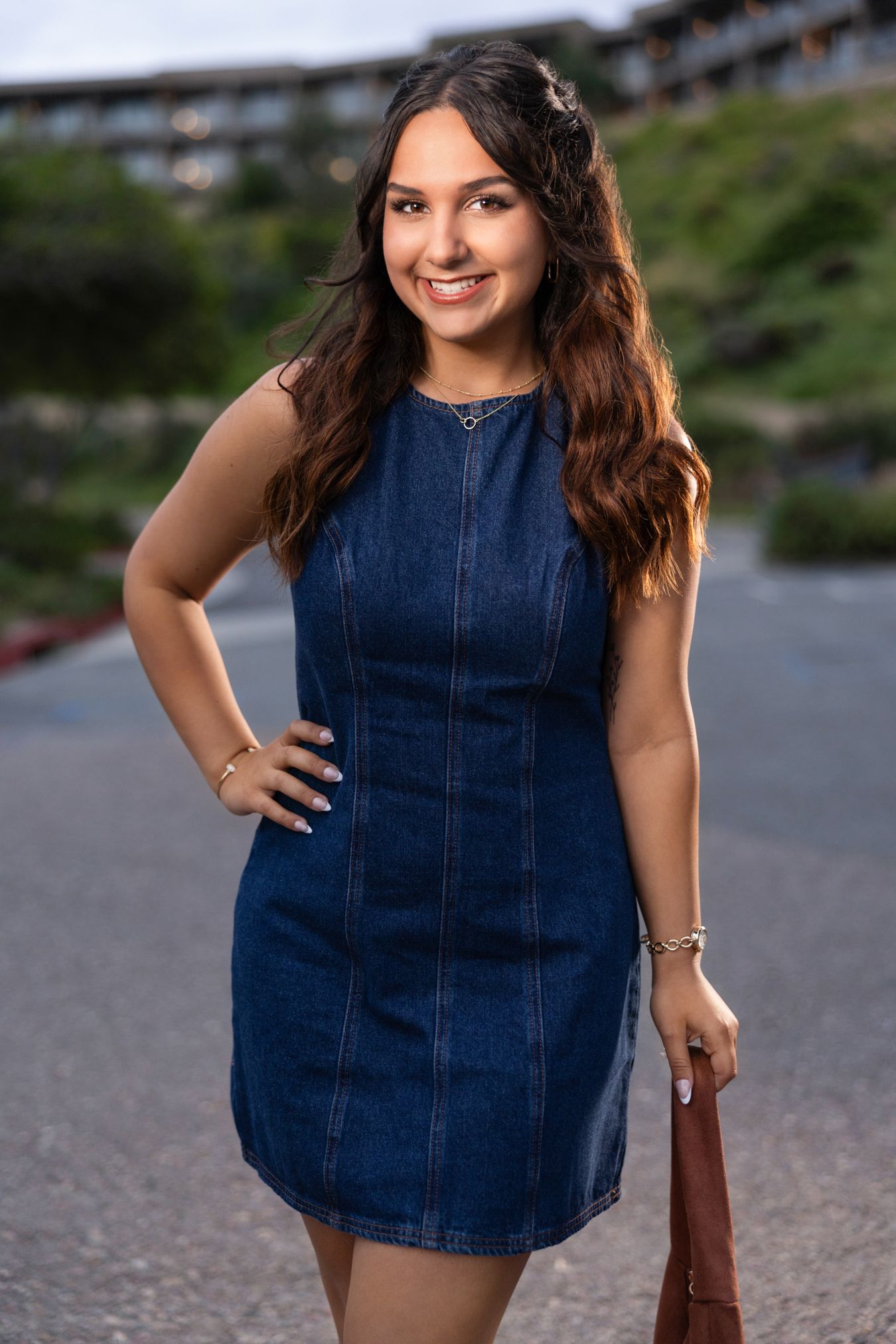 Young woman smiling in a blue denim mini dress, holding a brown bag, posing outdoors near a building.