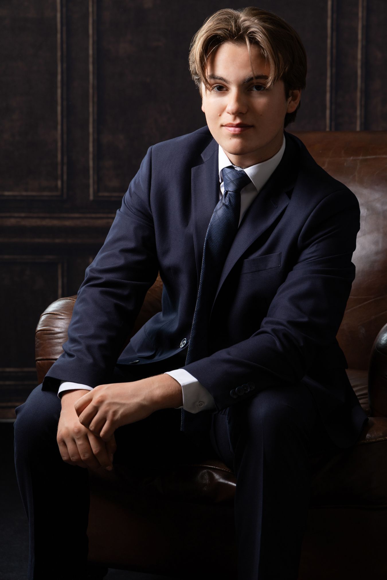 Young man in navy blue suit and tie sitting confidently in leather chair against dark wood paneled wall.