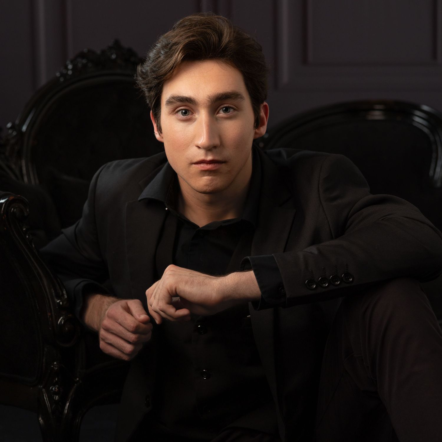 Young man in black suit sitting confidently in ornate dark chair against gray background.