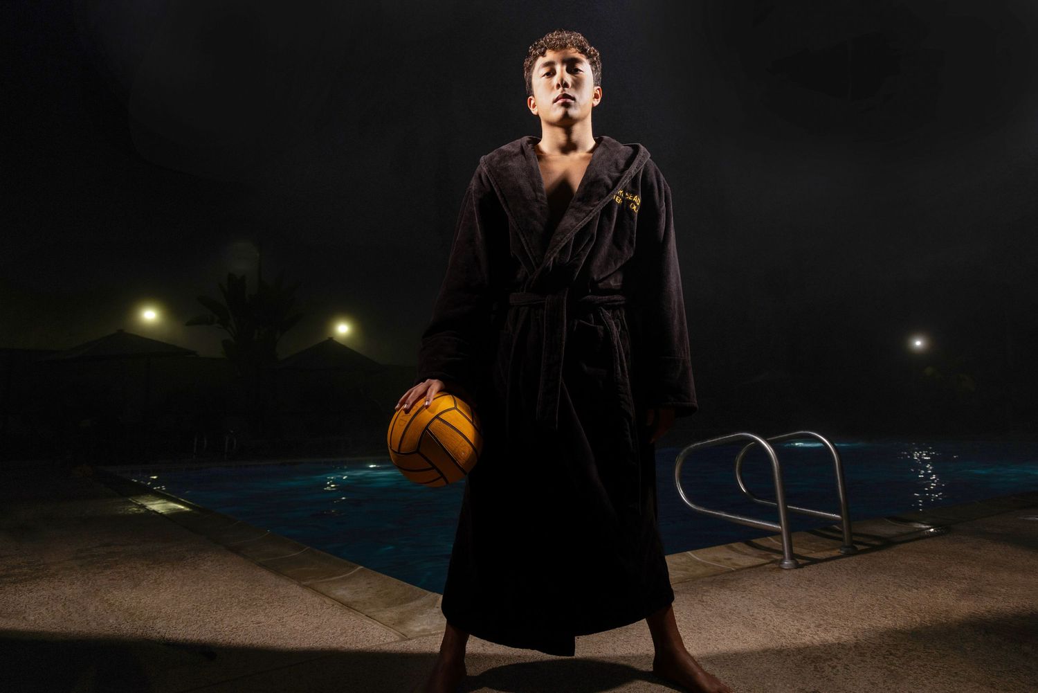 Athlete in black robe holds water polo ball poolside at night.