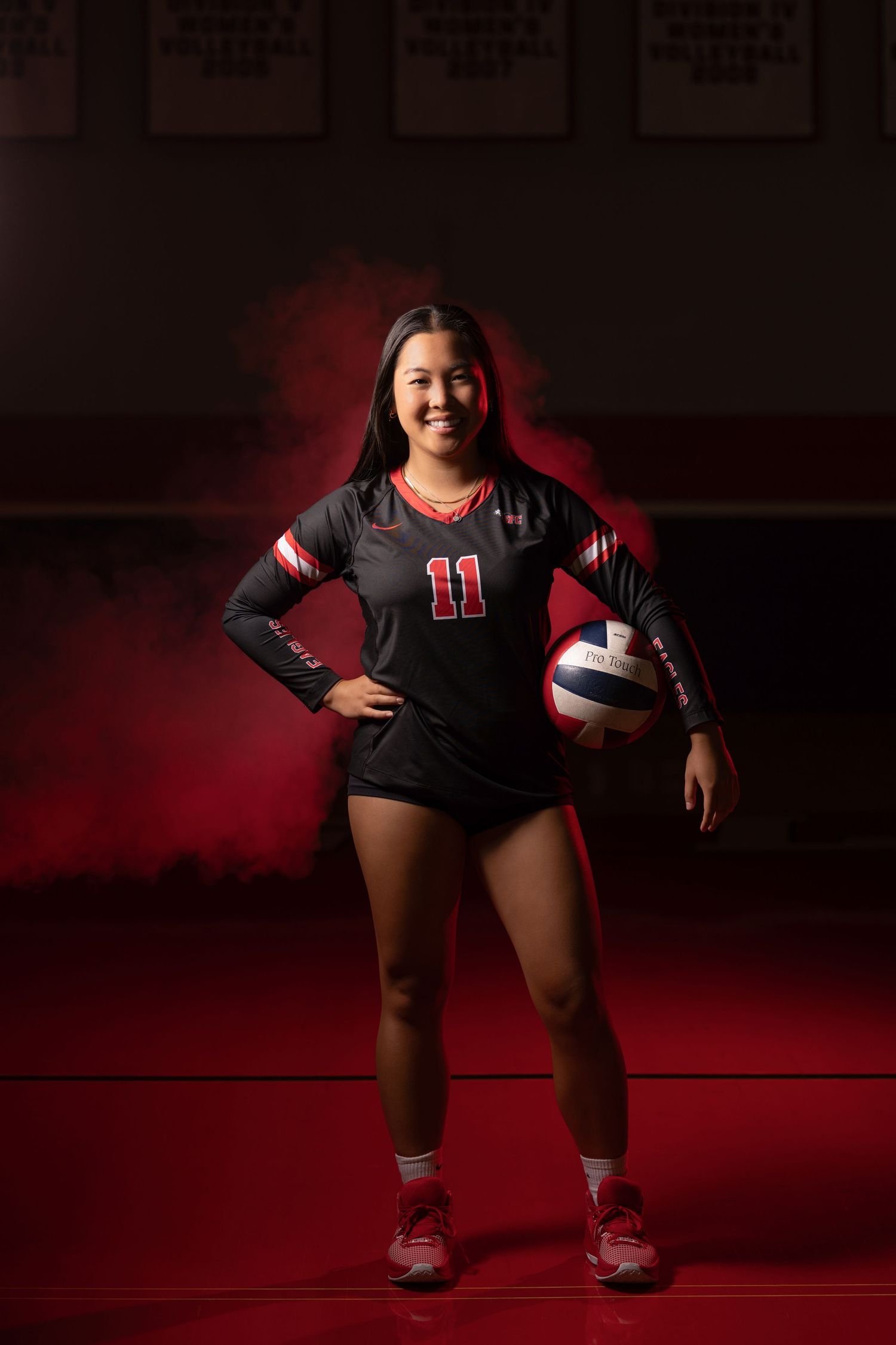 Smiling female volleyball player number 11 in black uniform posing with a ball in red-lit gymnasium.