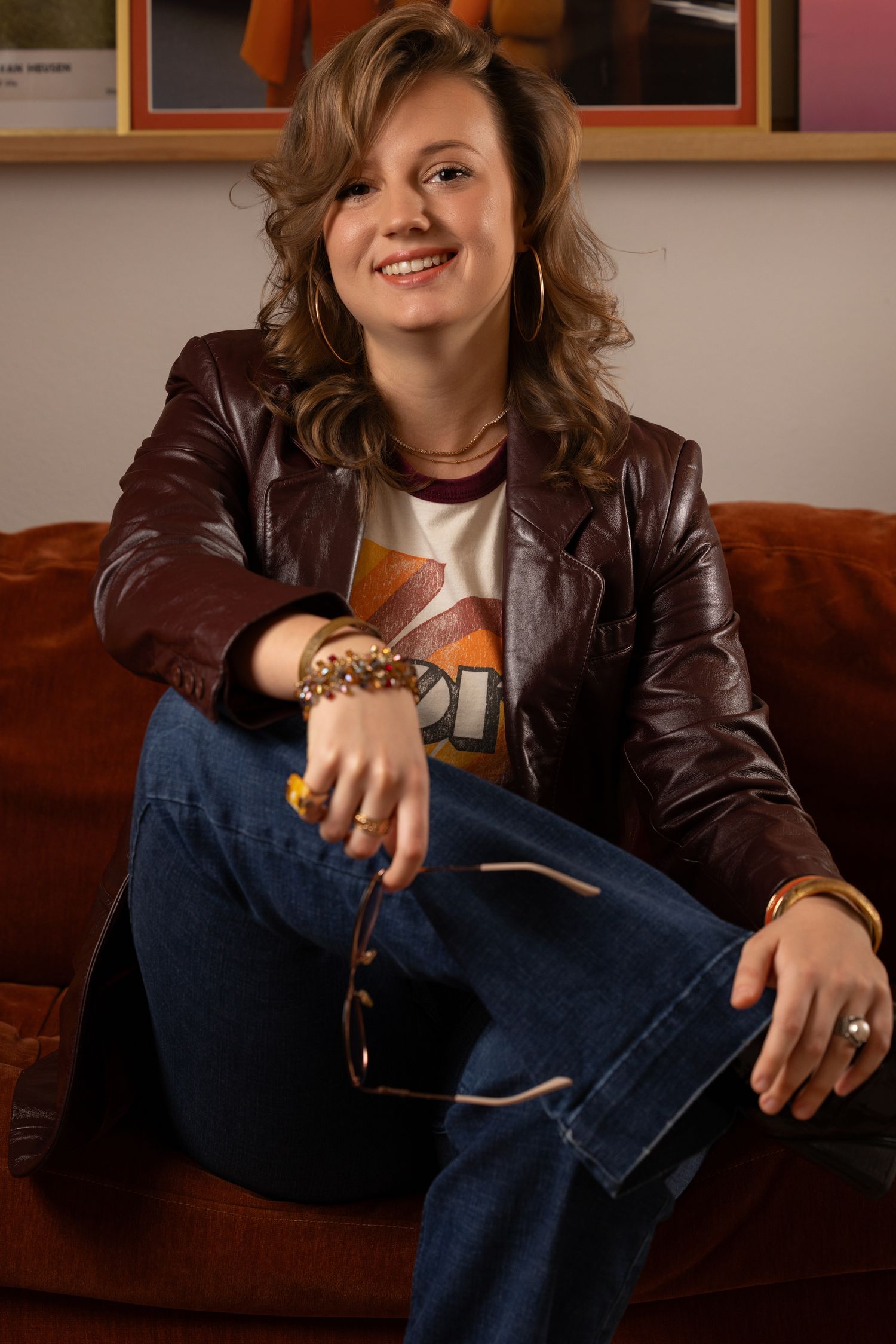 Smiling young woman in brown leather jacket and jeans sitting relaxed on an orange sofa wearing gold bracelets.