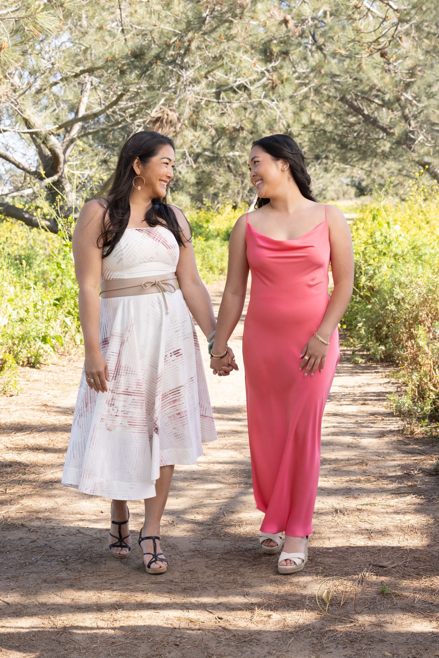 Two women in elegant dresses hold hands and smile at each other while walking on a sunny outdoor path.