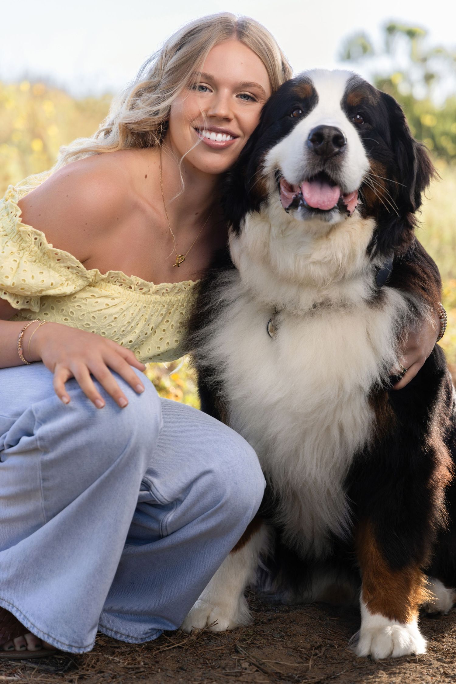 Smiling blonde woman in yellow top and jeans posing outdoors with a Bernese Mountain Dog.