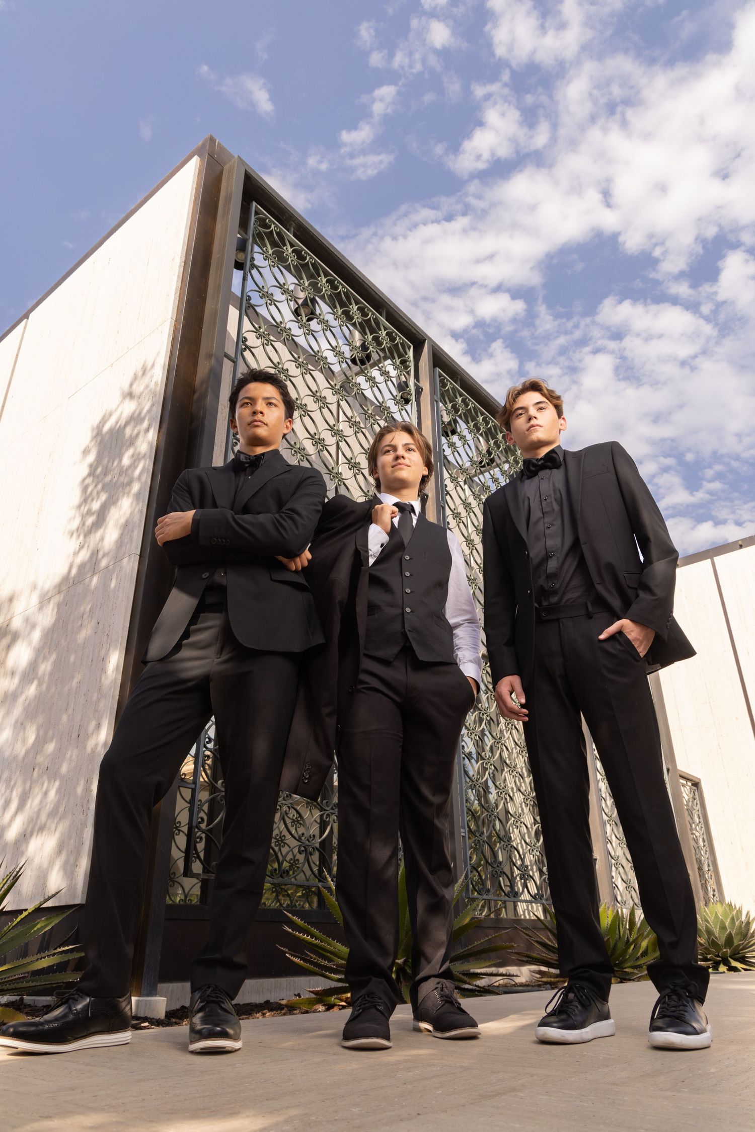 Three young men in formal black suits pose confidently outside a modern building under blue skies.