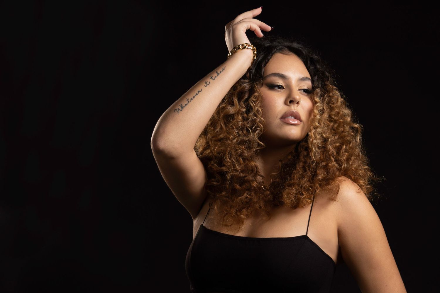 A young woman with curly auburn hair poses confidently in a black top against a dark studio background.