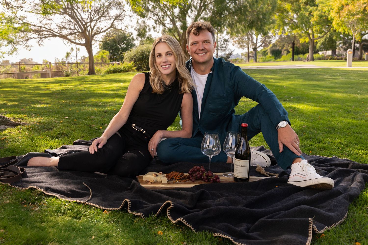 Couple smiling on a black picnic blanket with wine and charcuterie in a sunny park setting.