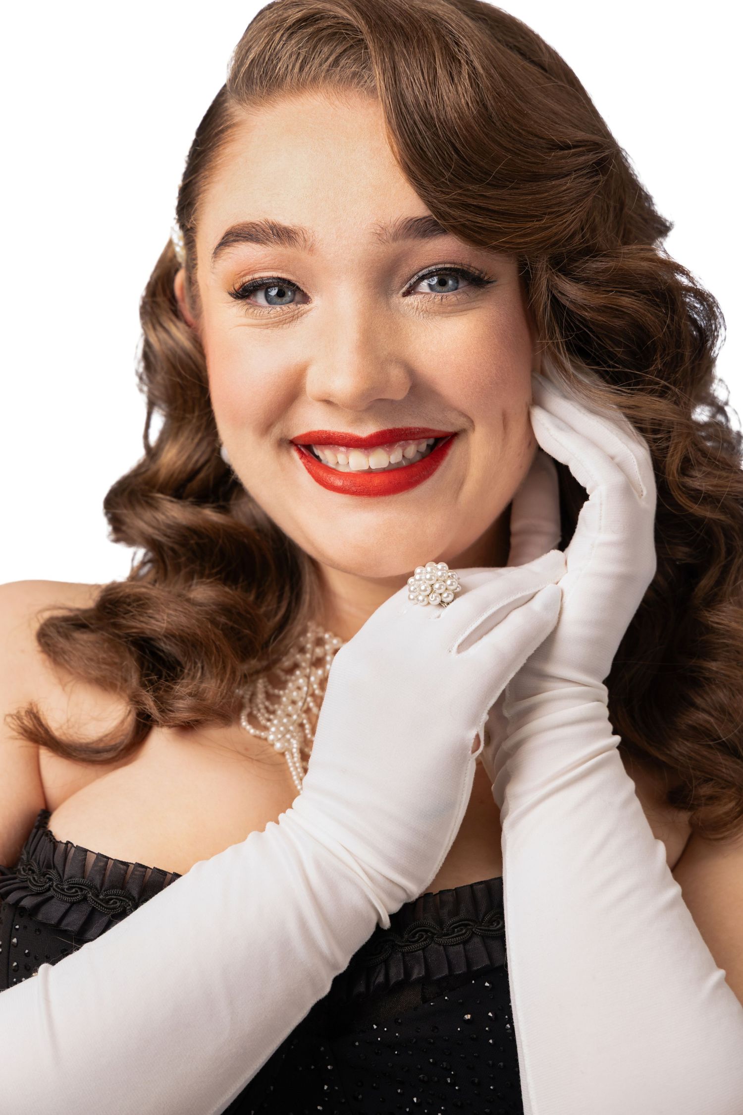 Elegant woman in vintage style with red lips, white gloves, pearl necklace, and curly hair poses gracefully.