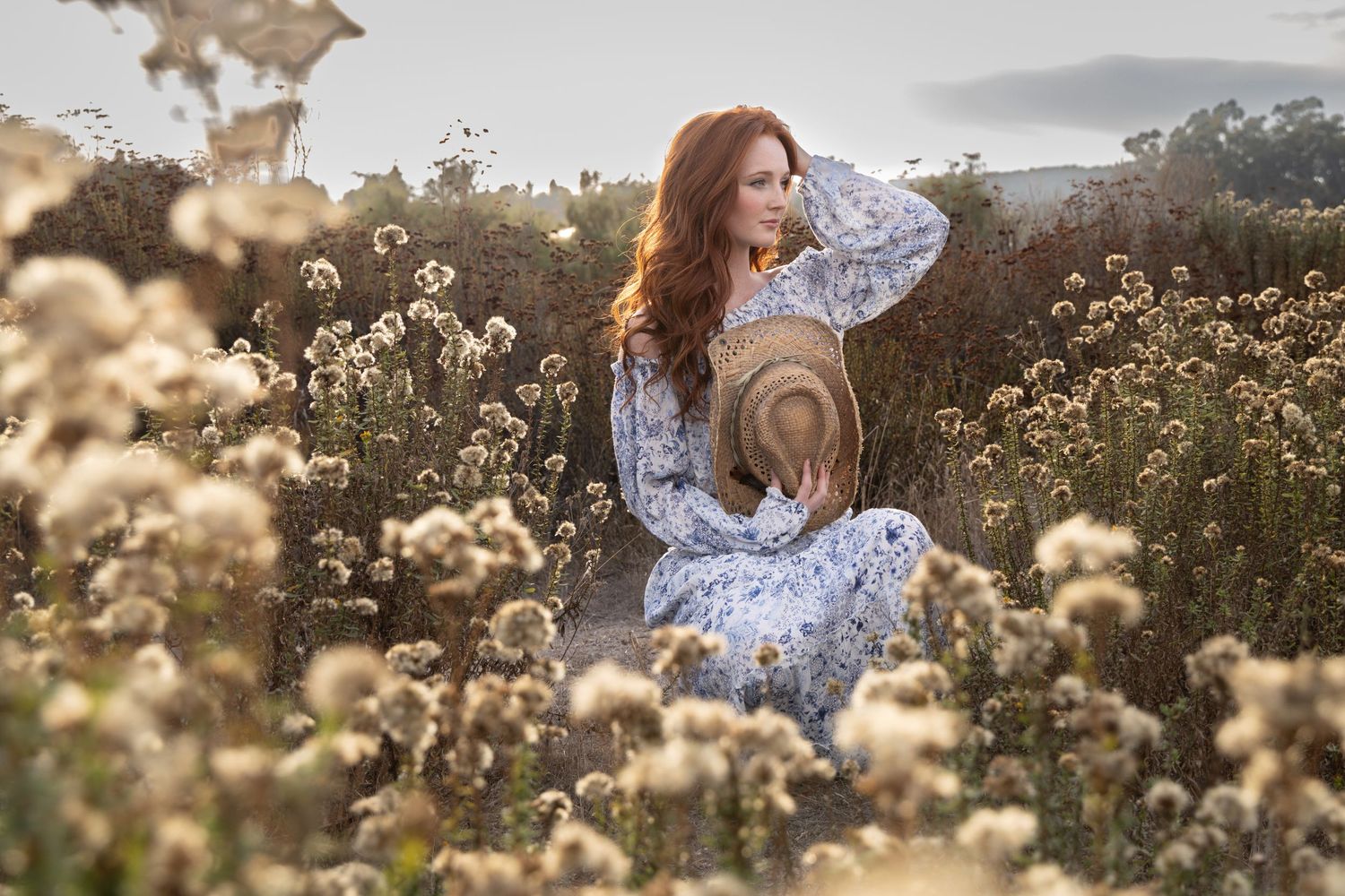 Redheaded woman in floral dress sits among wildflowers holding a straw hat.