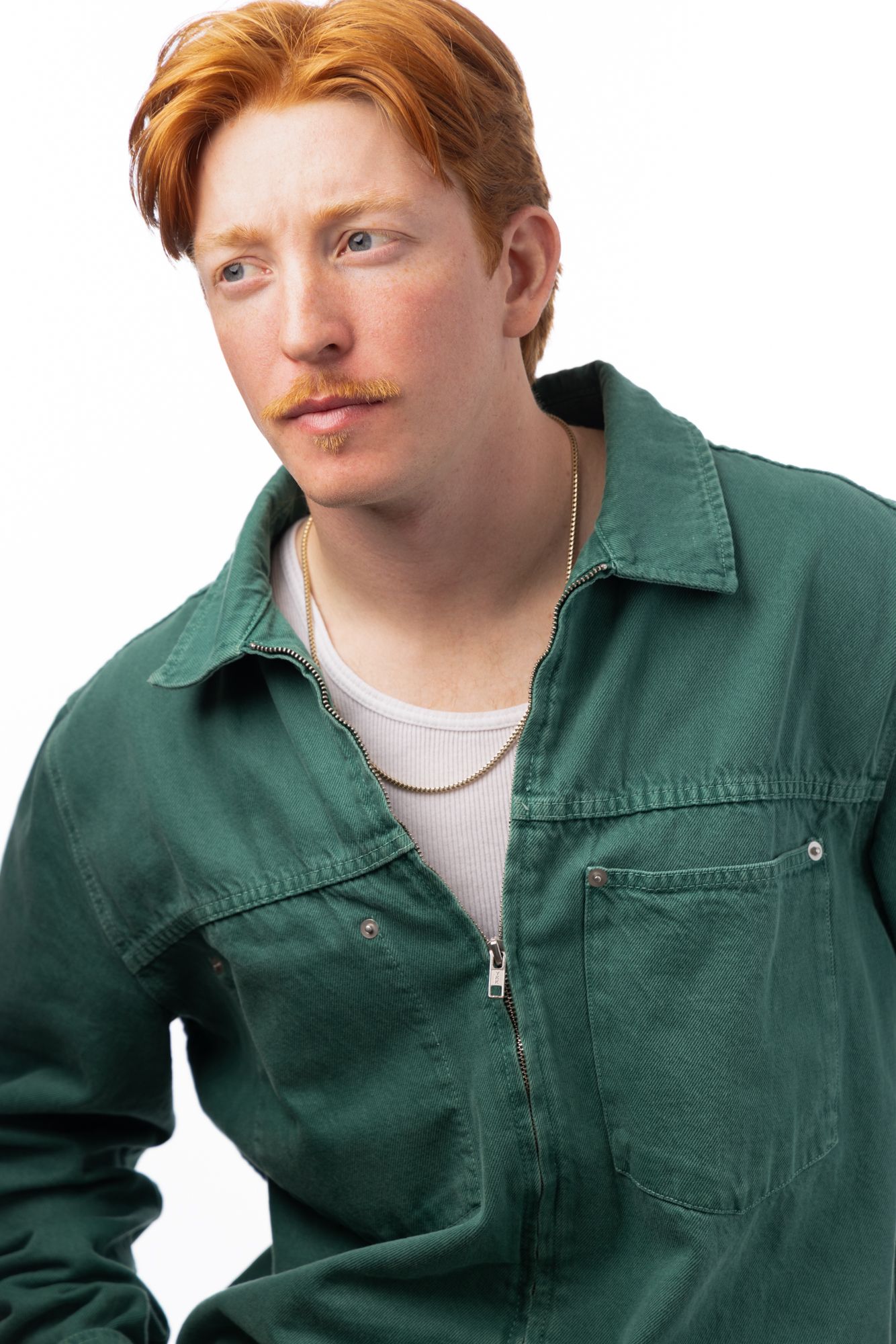 Young man with red hair and mustache wearing a green jacket and gold chain necklace against white background.