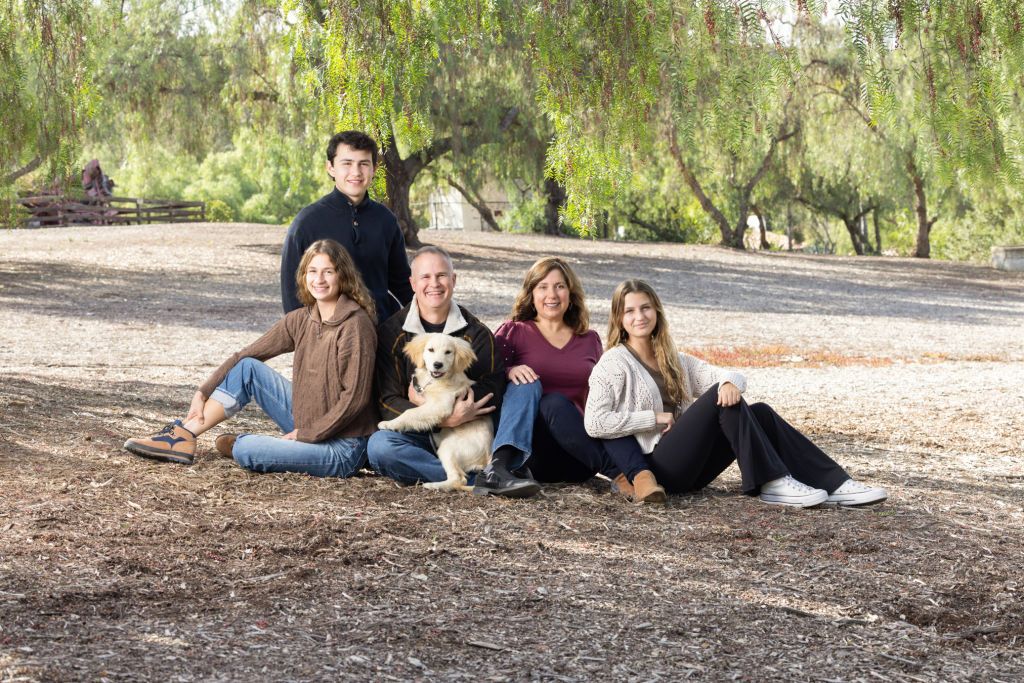 Family of five with a golden retriever dog sitting outdoors under willow trees on a sunny day.