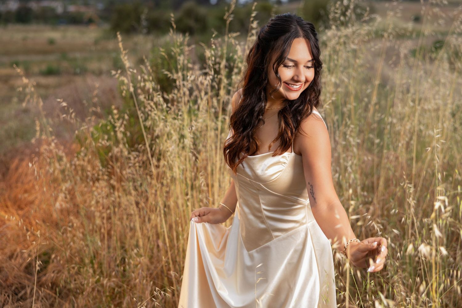 A smiling young woman in a flowing white satin dress stands gracefully in a golden field of tall grass.