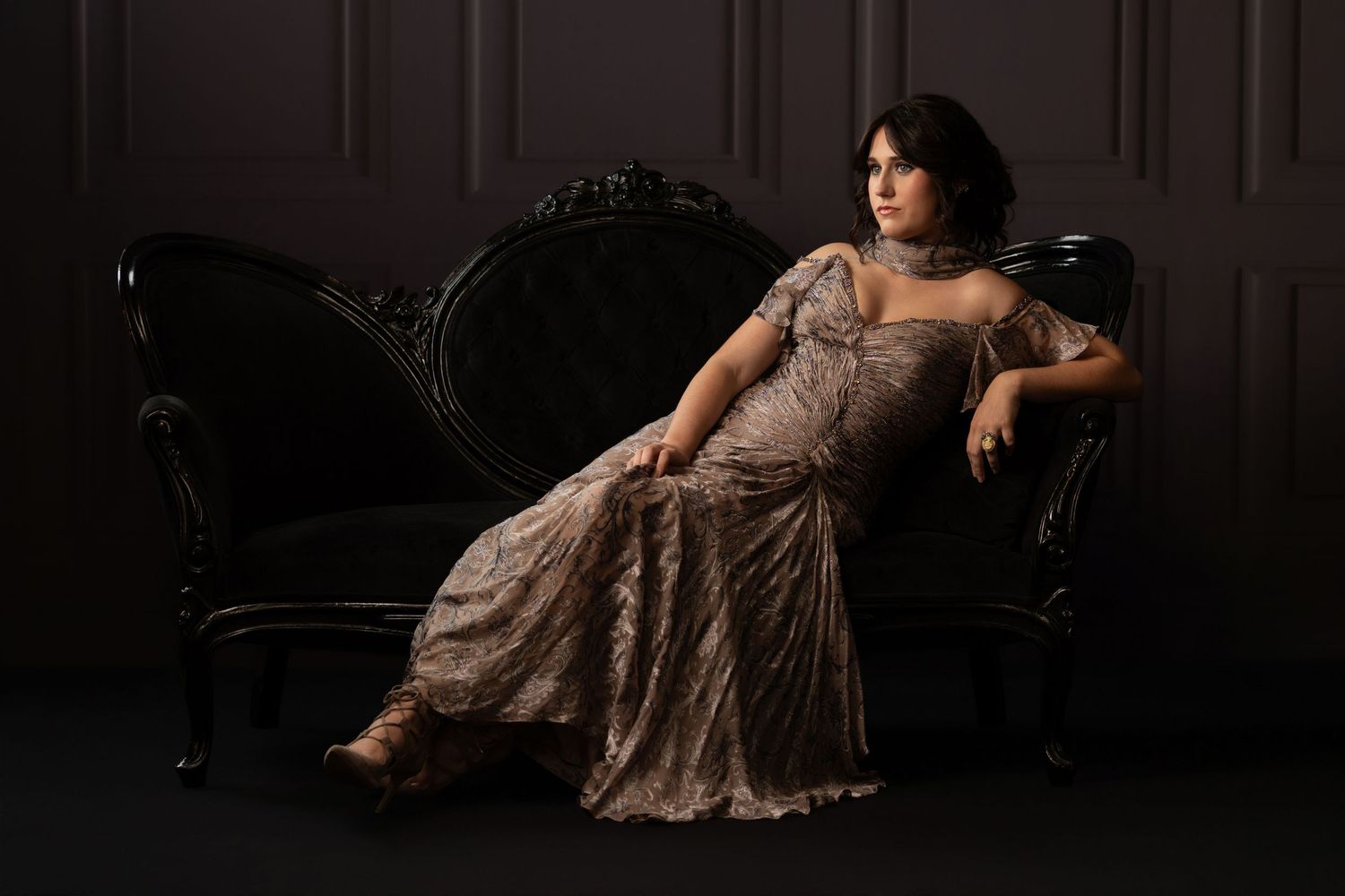 Woman in elegant lace gown reclining gracefully on ornate vintage chaise lounge in dramatic dark studio setting.