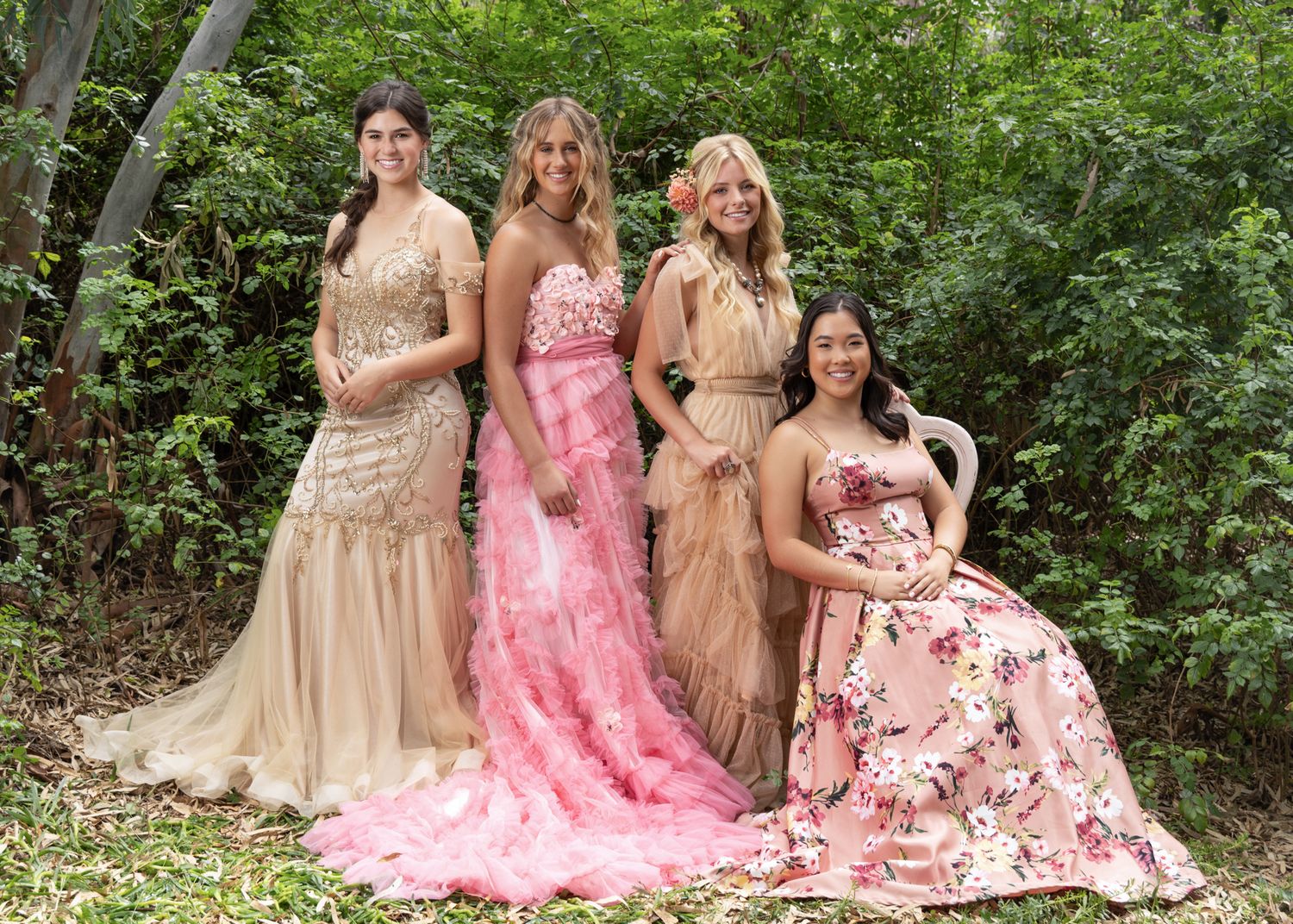 Four women in elegant formal gowns pose together outdoors among lush green bushes.