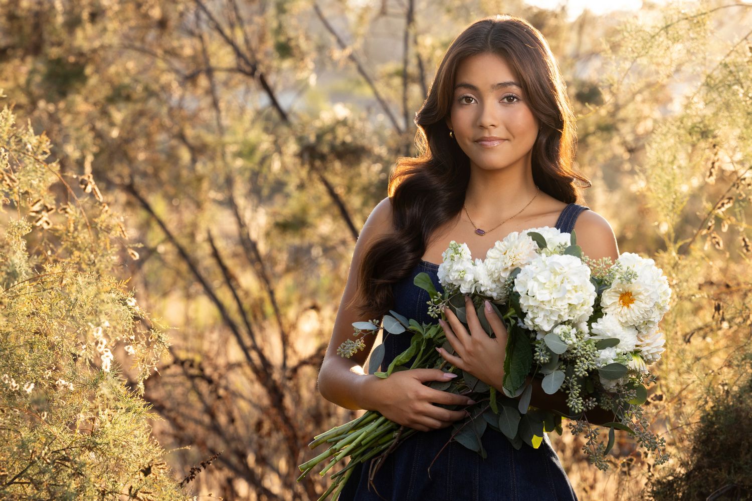 Young woman in navy dress holding white flower bouquet outdoors in golden light.