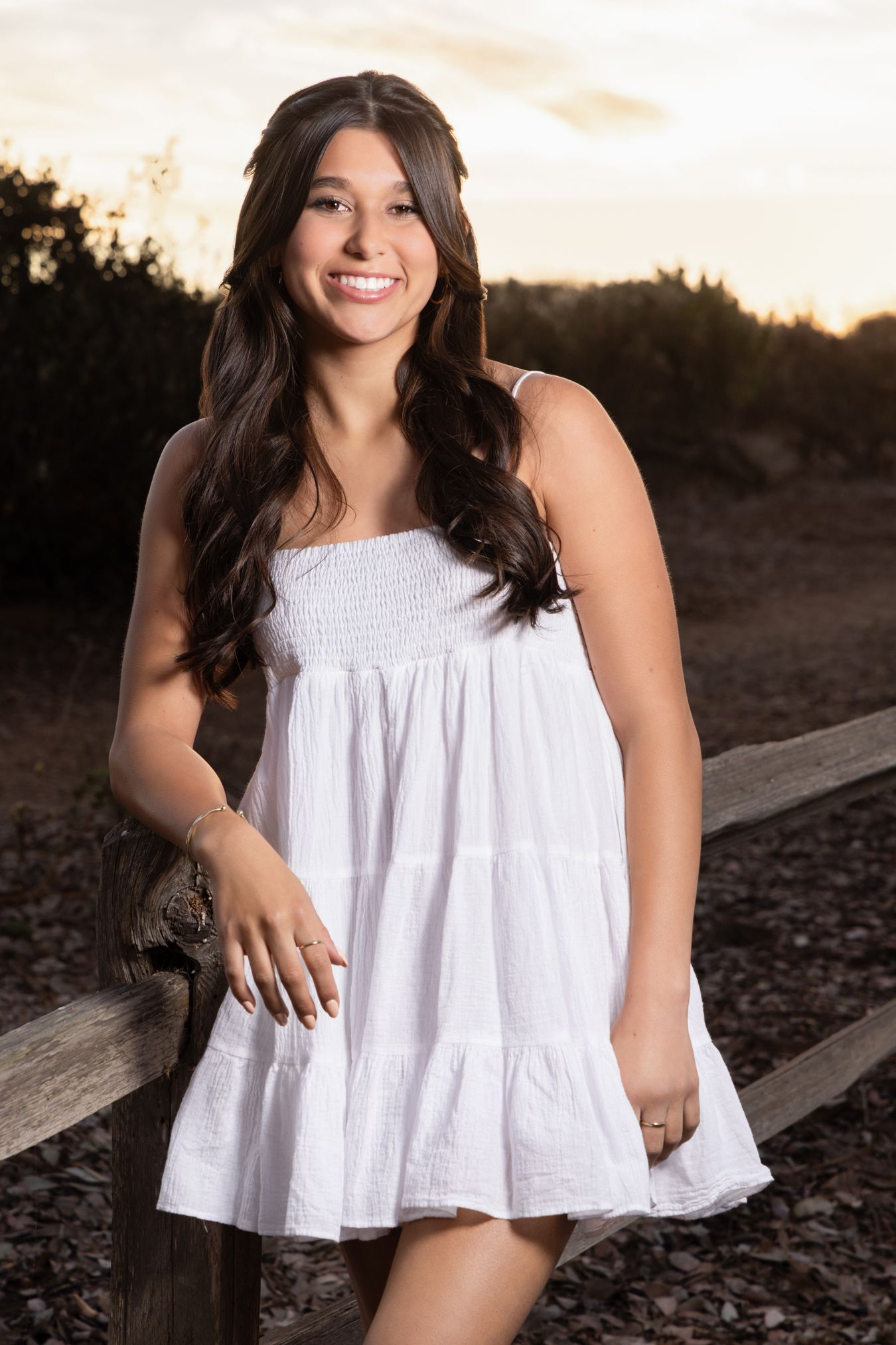 Smiling young brunette woman in white sundress leaning on wooden fence at golden hour sunset.