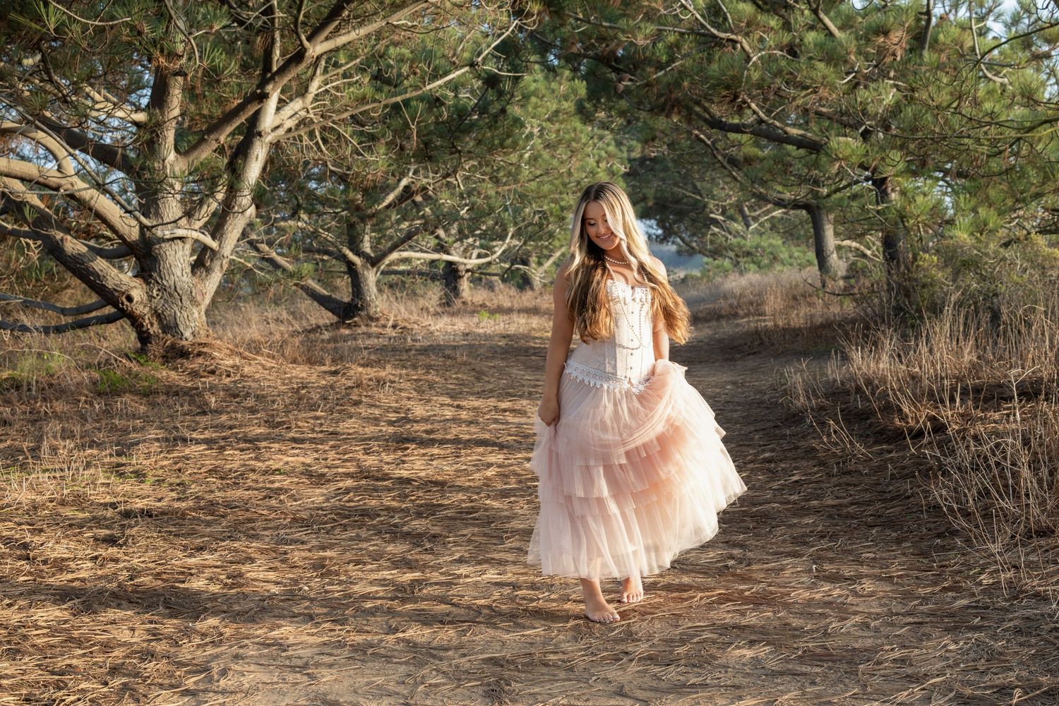 A young girl in a pink tulle dress walks along a sandy forest path surrounded by pine trees.