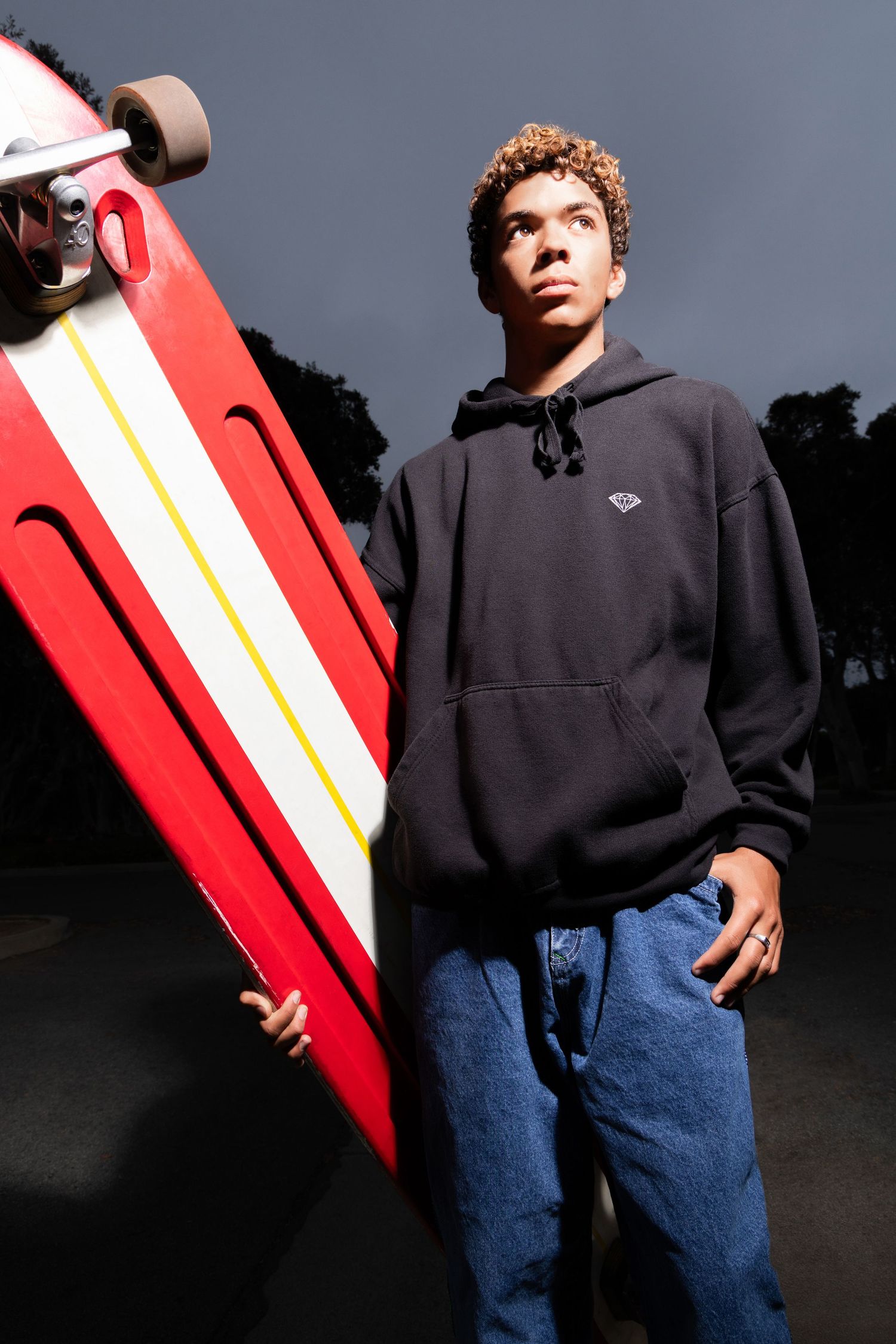 Young skater holding a red and white longboard, wearing a black hoodie and jeans outdoors at dusk.