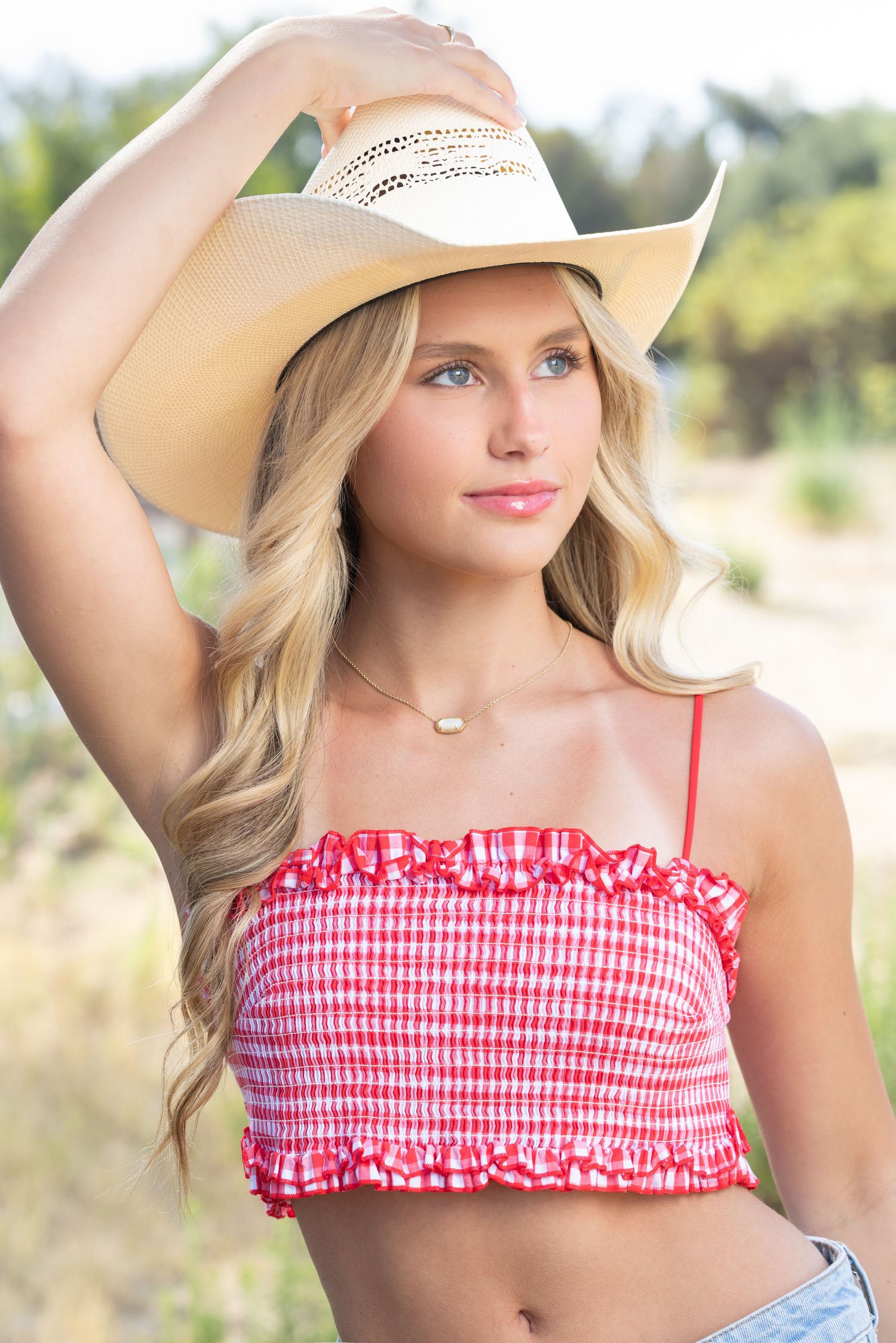 Young blonde woman in red gingham crop top and white cowboy hat posing outdoors in a sunny field.