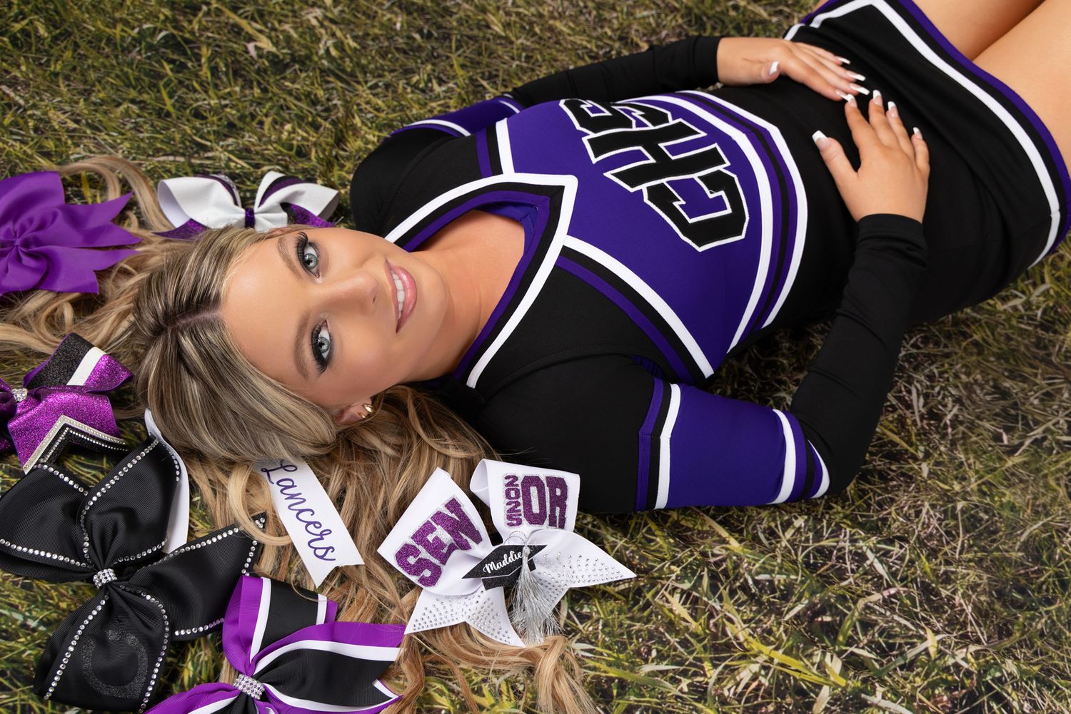 Cheerleader in purple and black uniform poses lying on grass field.