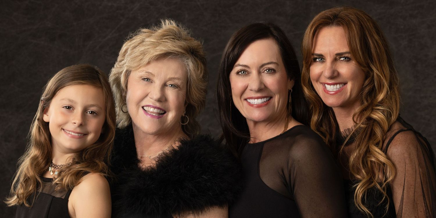 Four generations of women in black outfits smiling together in a professional studio portrait photograph.