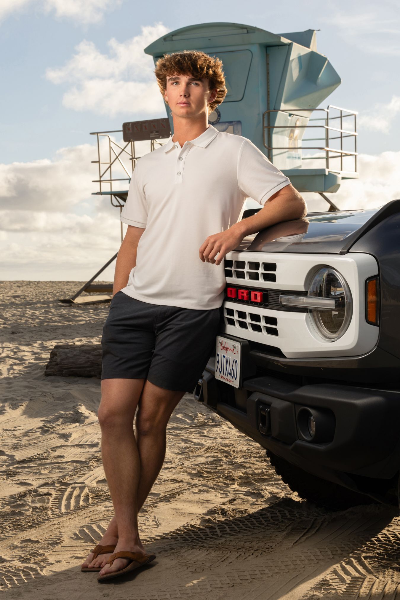 Young man in white polo and shorts leaning on a Ford Bronco on a sandy beach near a lifeguard tower.