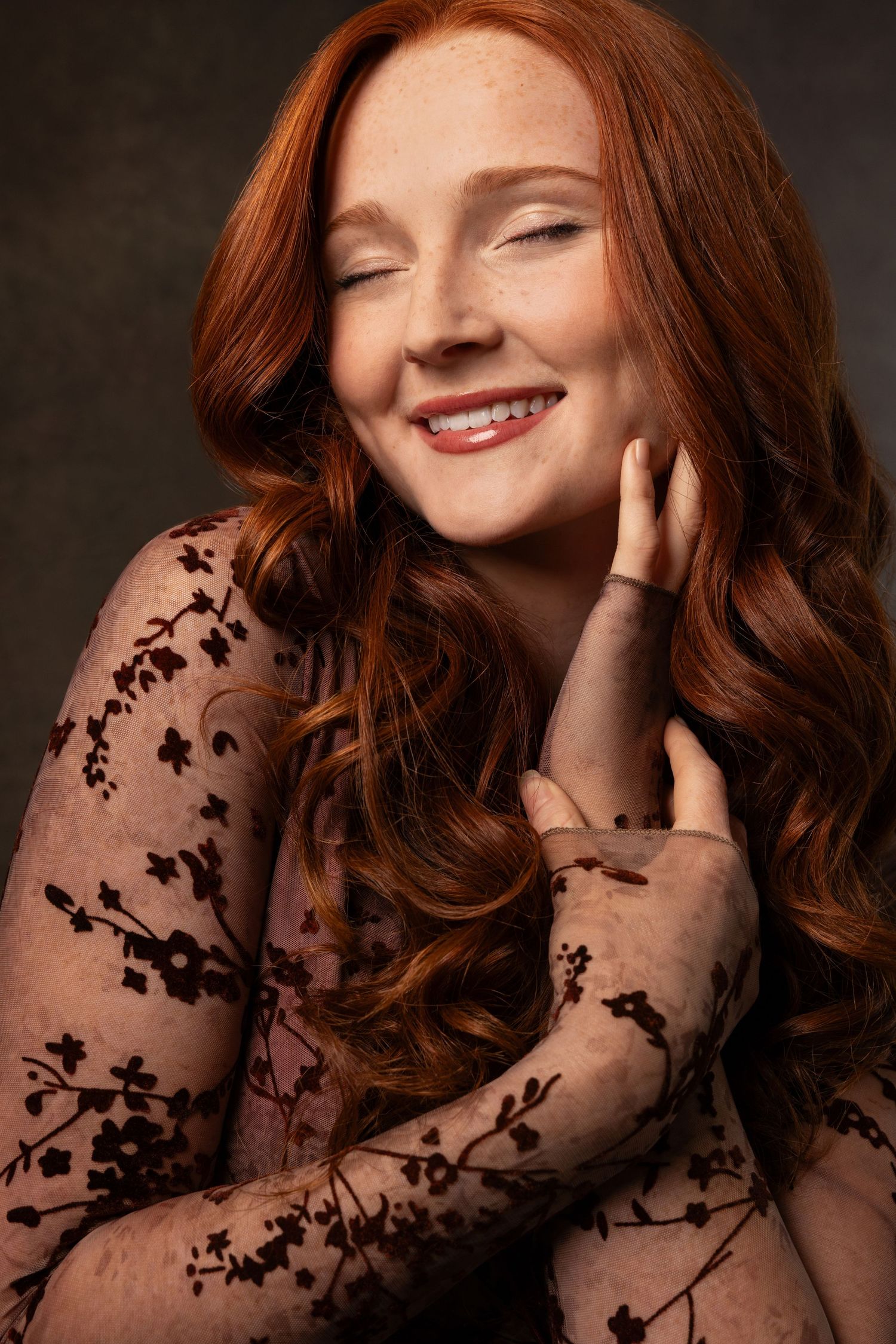 A smiling redhead woman with freckles in a floral lace dress, eyes closed, touching her face against a dark background.