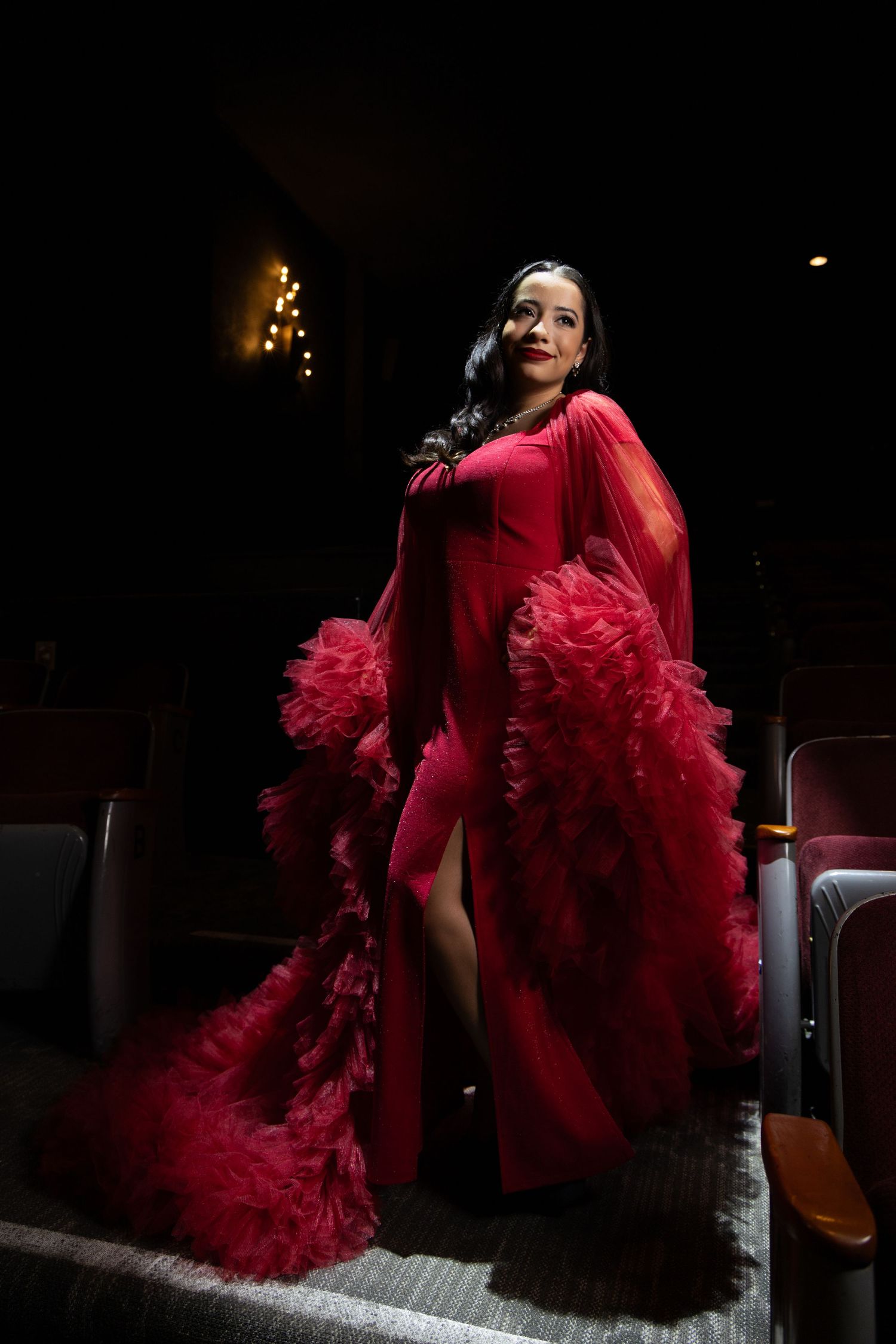 Woman in elegant red ruffled gown poses dramatically in a dimly lit theater aisle.