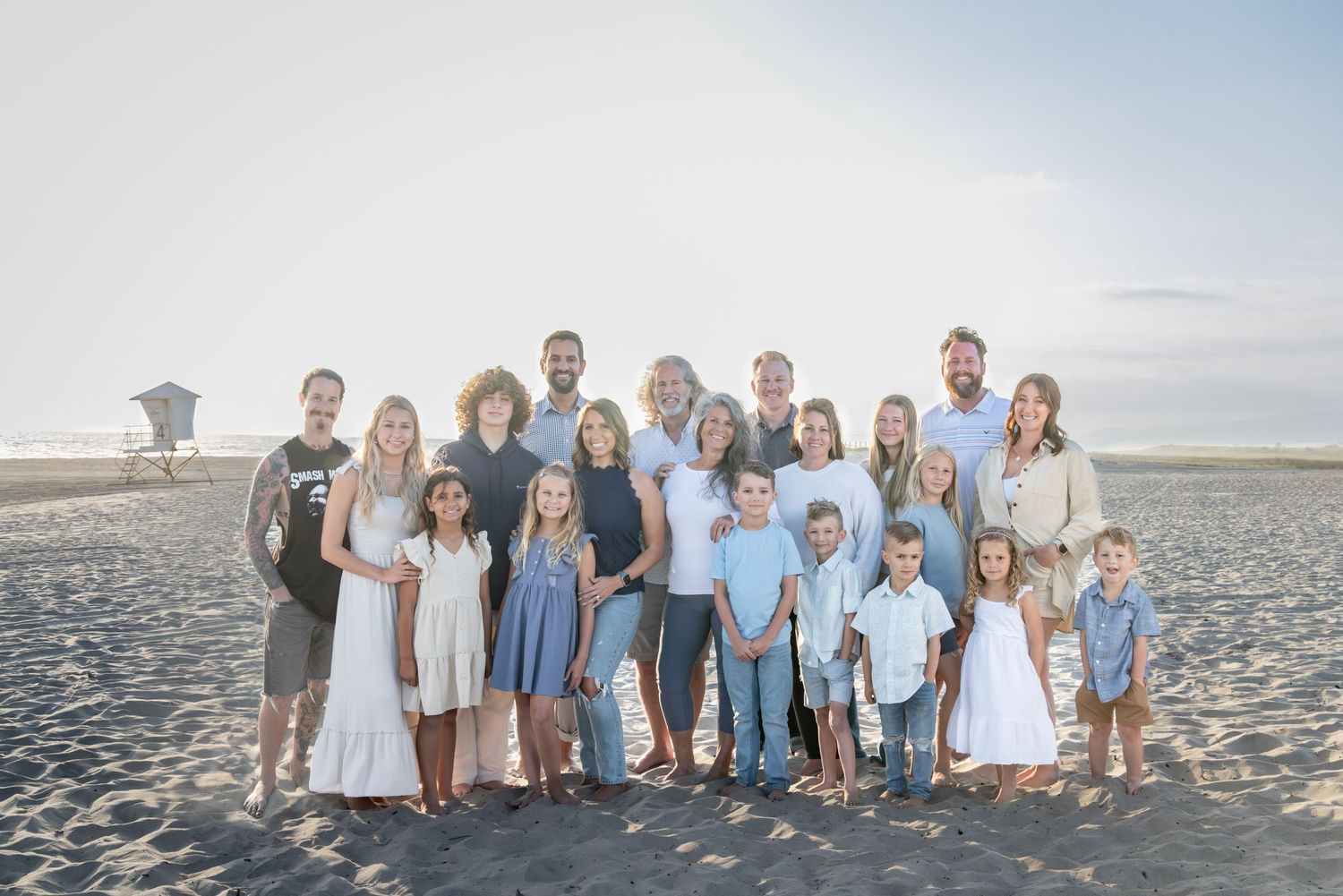 Large family group portrait on a sandy beach, smiling together in casual light-colored outfits on a sunny day.