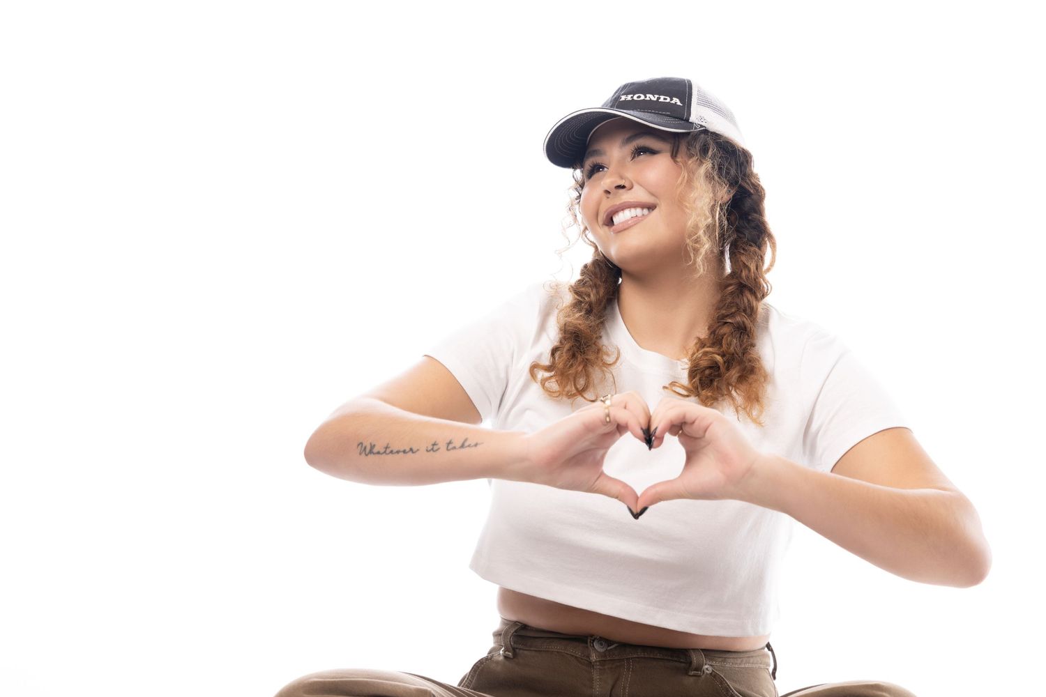 Smiling young woman in white crop top and black cap making a heart shape with her hands.