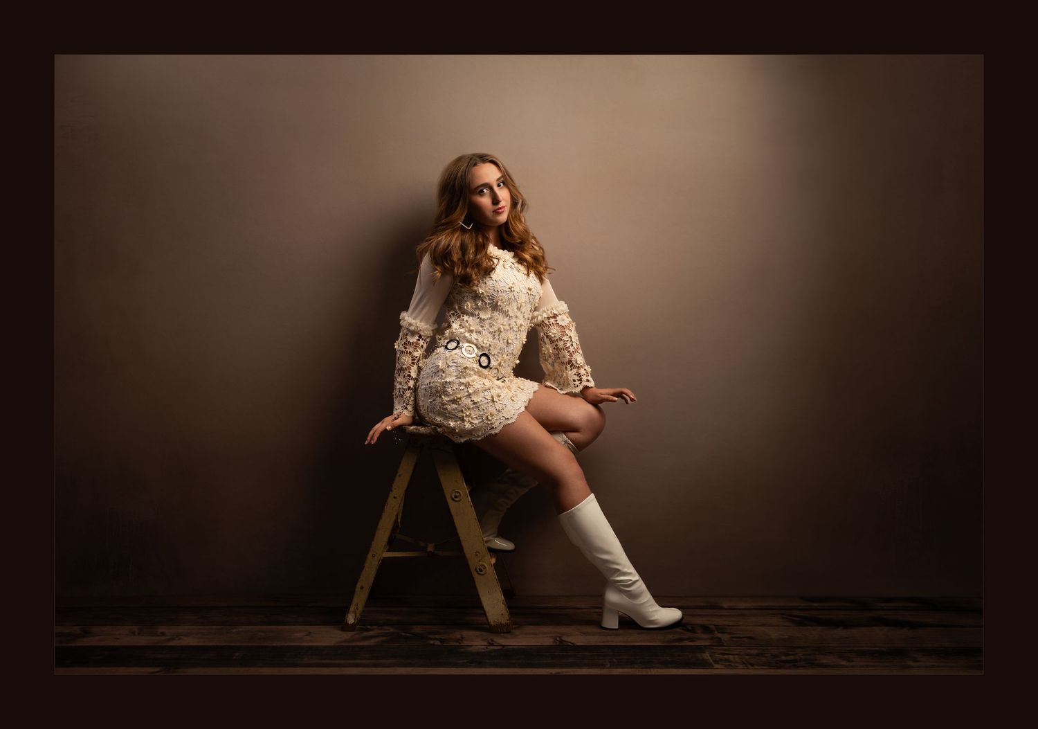 Young woman in cream lace mini dress and white knee-high boots seated on wooden stool against brown backdrop.