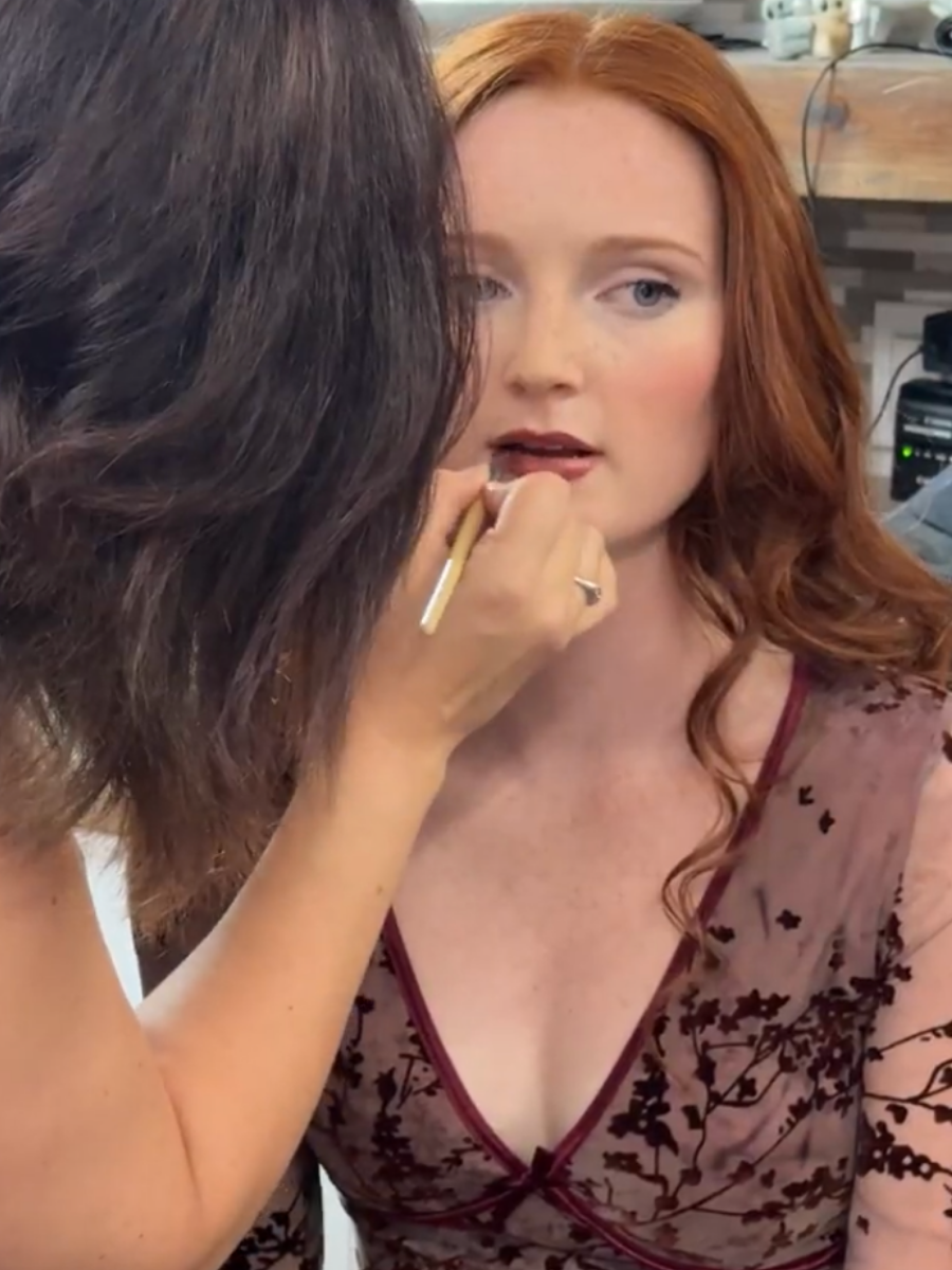 Makeup artist applying dark lipstick to redhead woman in floral lace top.