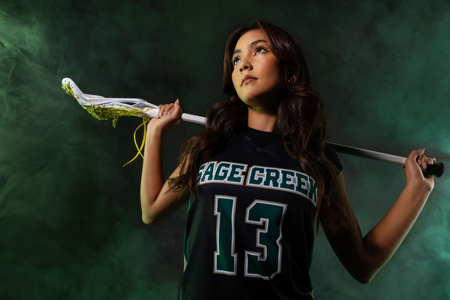 Female lacrosse player in Sage Creek number 13 jersey holding stick against dramatic green smoky background.
