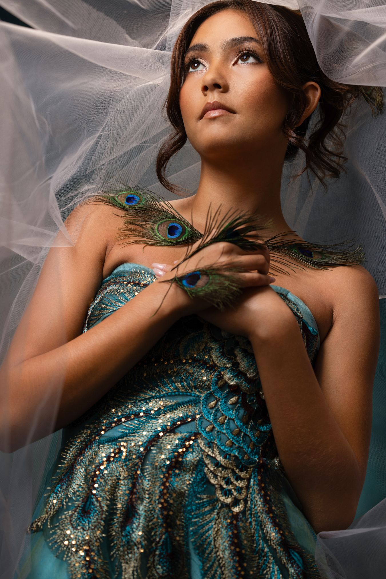 Woman in teal beaded gown holding peacock feathers under sheer veil in studio.