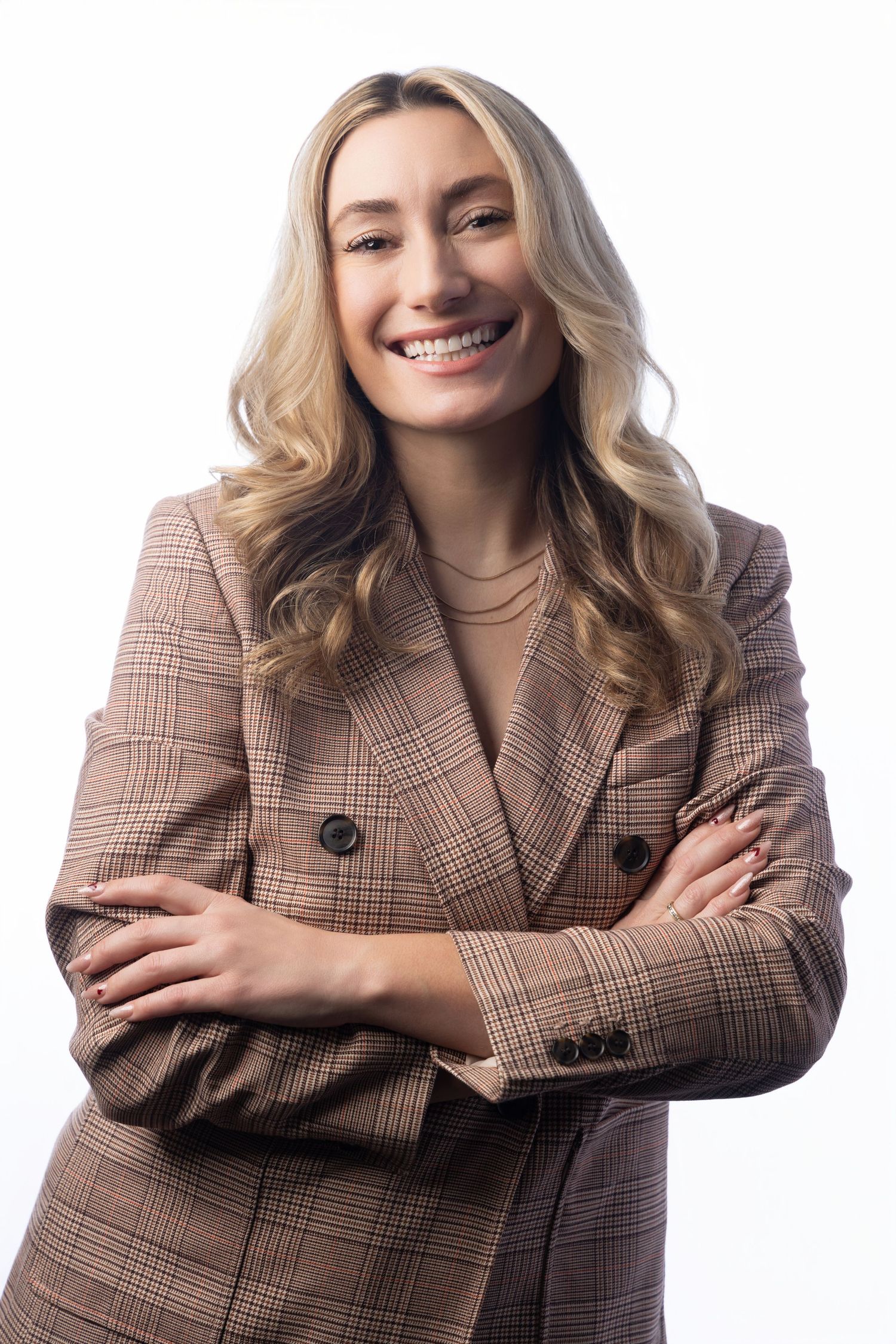 Smiling blonde woman in a brown plaid blazer with arms crossed against a white background.